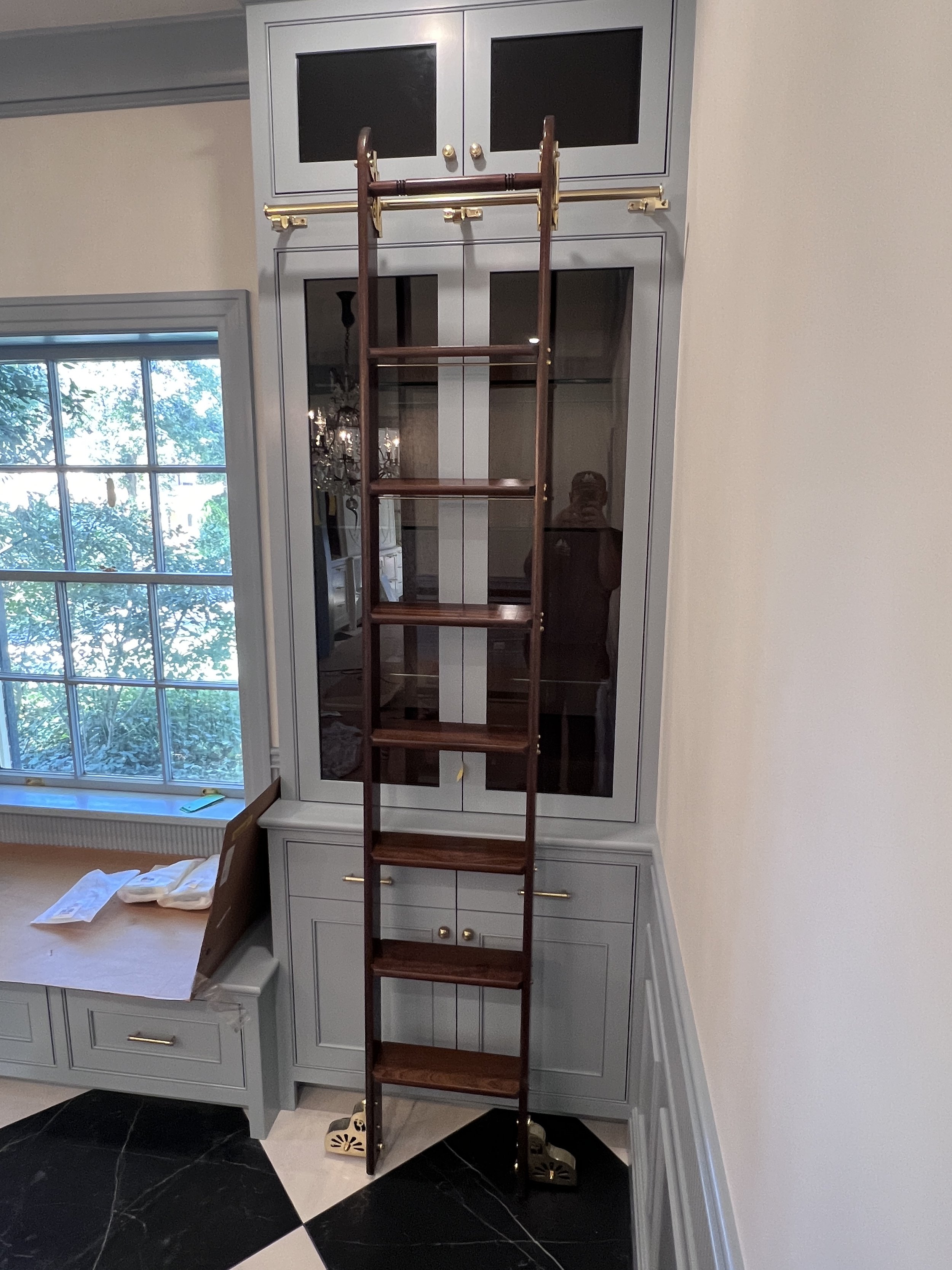 A wooden ladder positioned in front of a built-in cabinet with glass doors in a room with a large window showing trees outside.