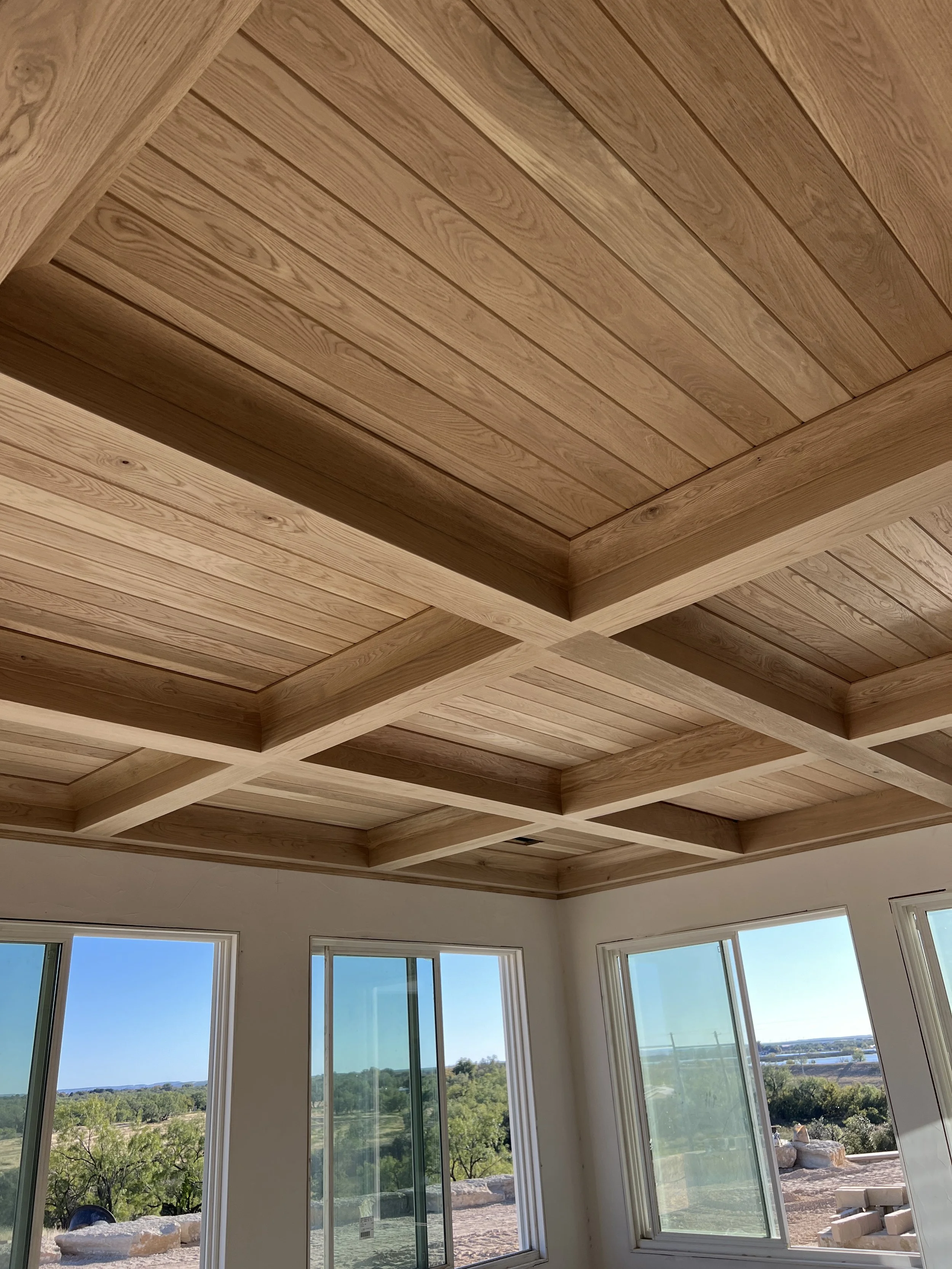 Interior view of a room with a wooden coffered ceiling and large windows revealing a scenic outdoor landscape with trees and hills.
