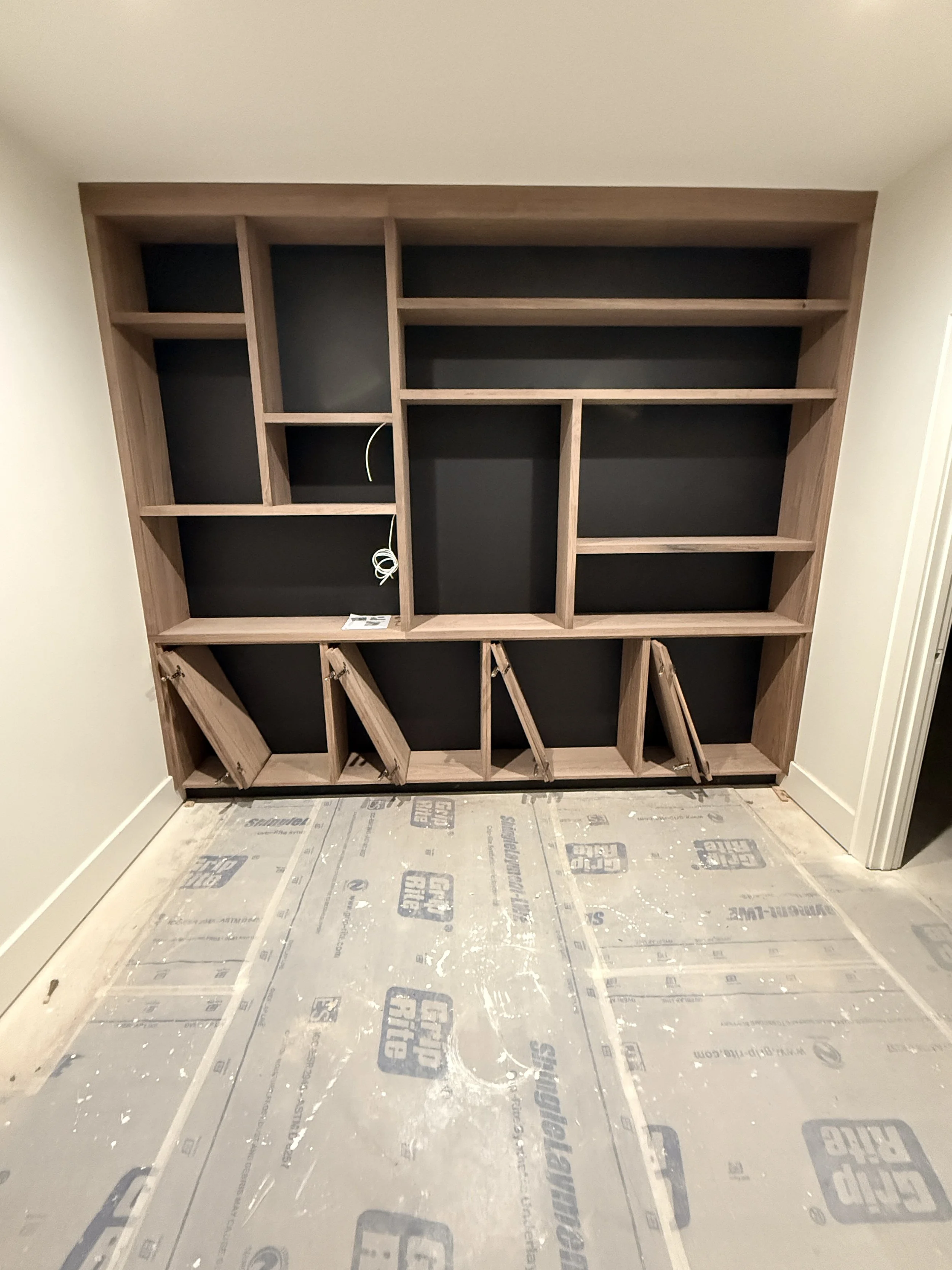 Empty built-in wooden bookshelf with black backing, partially installed on an unfinished room with drywall walls and a floor covered in protective paper.