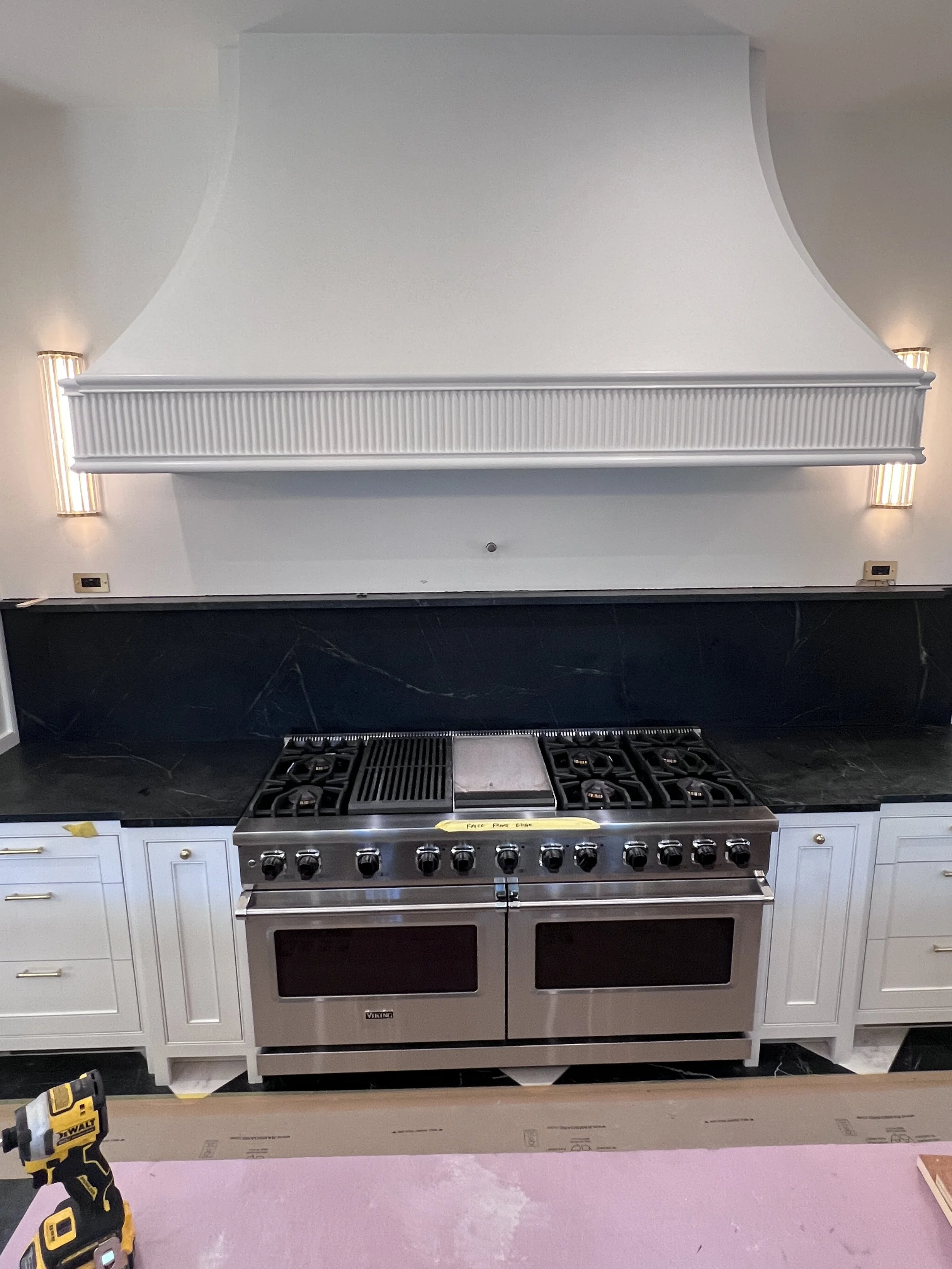 A kitchen with a black countertop and white cabinets. A stainless steel Viking oven and stove with black knobs, and a white range hood above it, which is still being installed. There are two wall-mounted lights on either side of the range hood and el