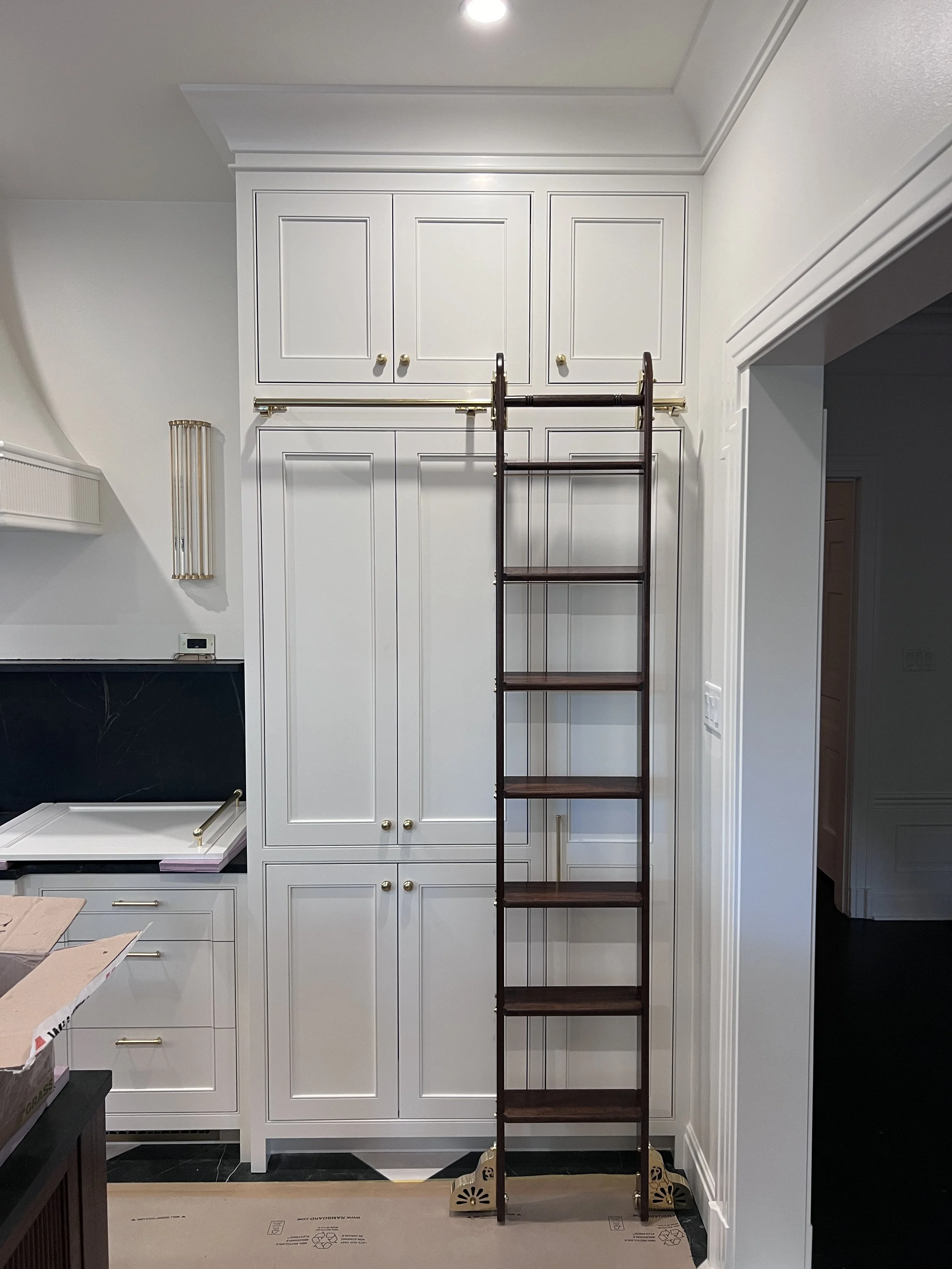 A tall wooden ladder with five steps is positioned in front of white built-in cabinets with gold knobs, in a room with white walls and crown molding. A dark marble fireplace is partially visible on the left, and part of a cardboard box is visible on 