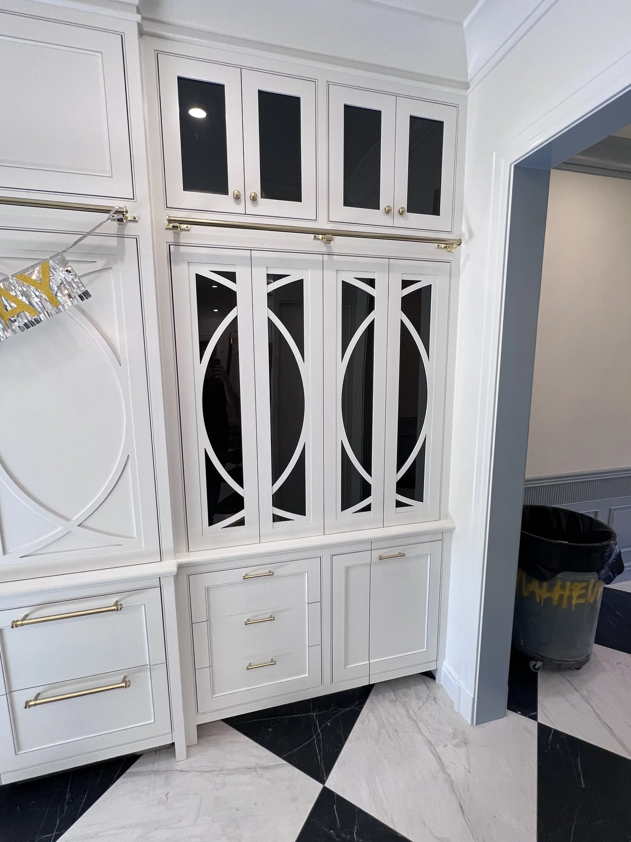 White built-in cabinet with glass-paned upper doors and gold handles, situated next to a grey wall and a trash can with a yellow and black bag inside.