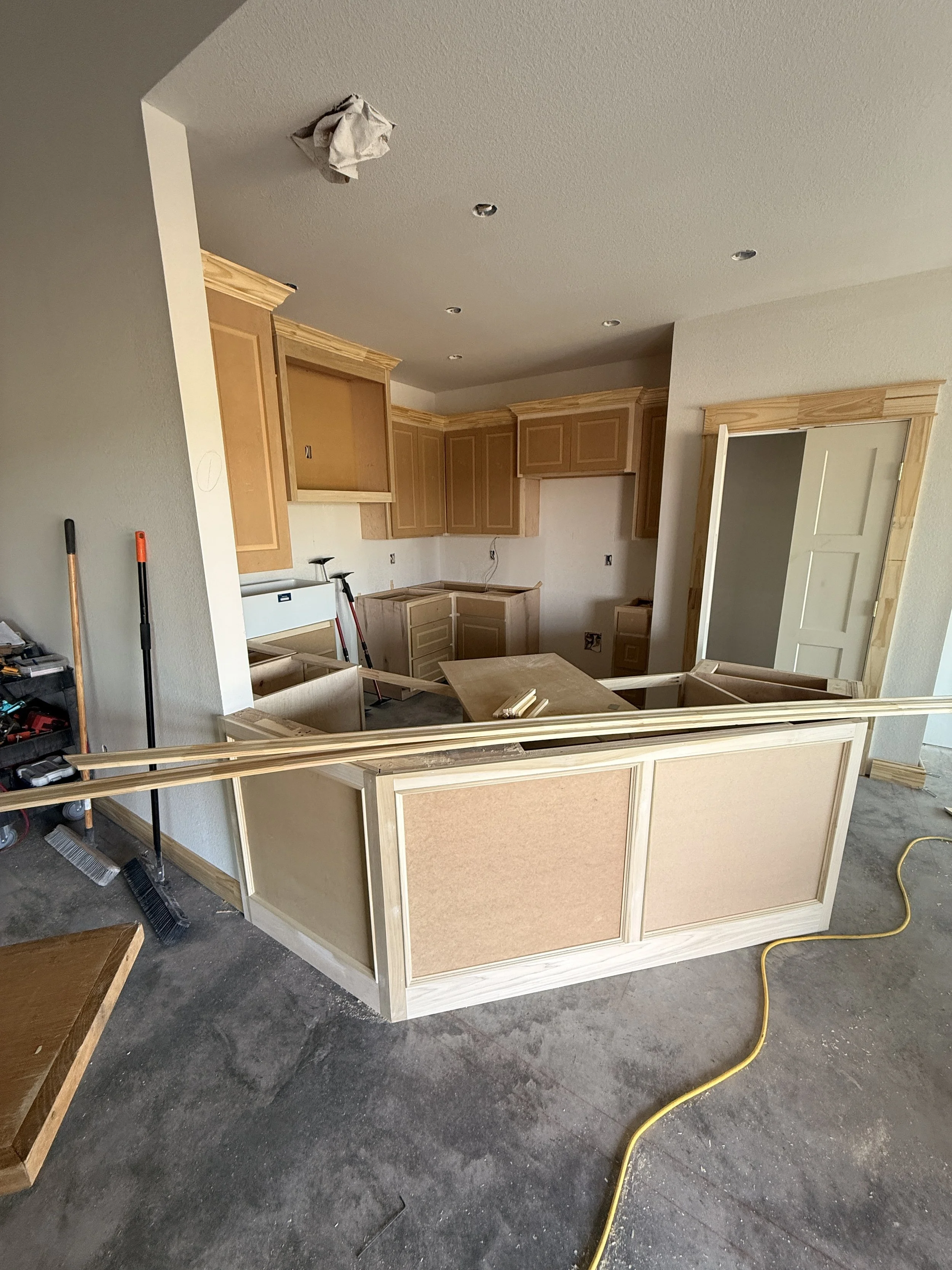 Interior of a kitchen under construction with unfinished wooden cabinets, a partially installed door frame, and tools and construction materials on the floor.