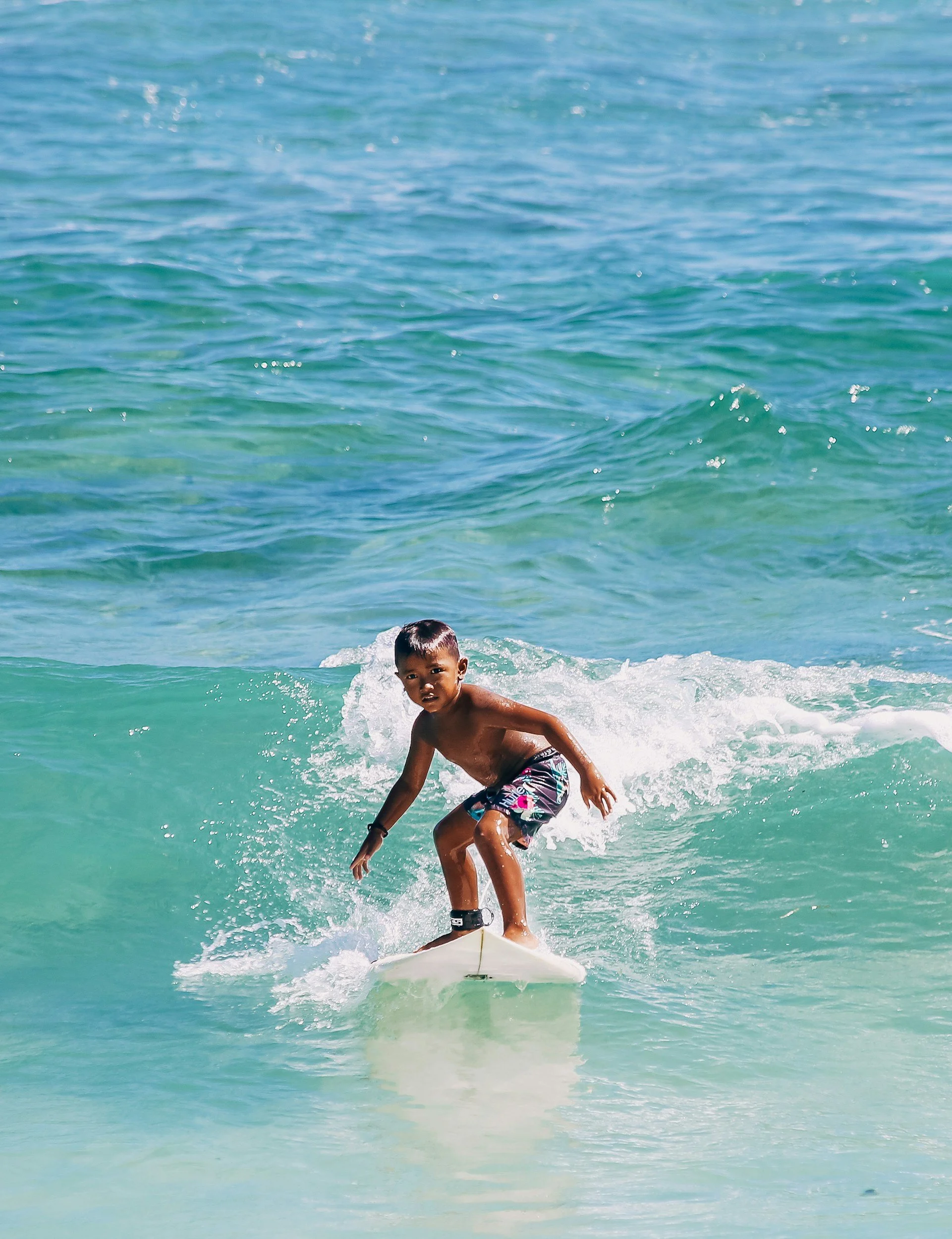 A young boy surfing on a surfboard in the ocean, with the turquoise water and waves around him.