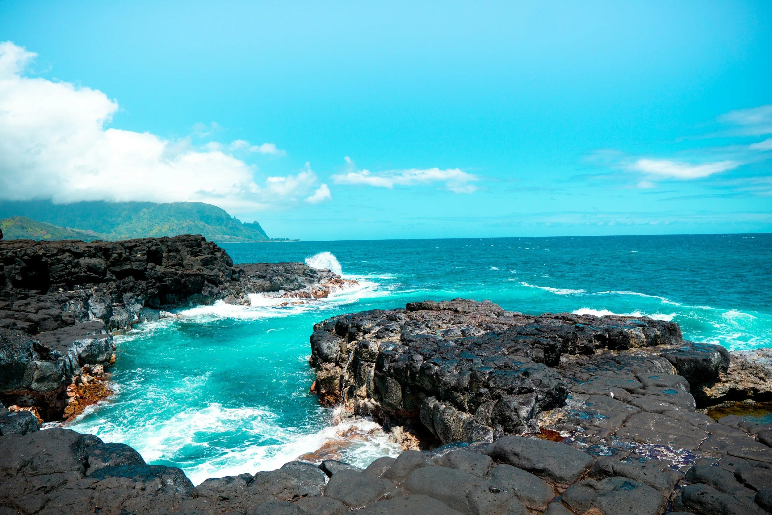 Photo of a rocky coastline with turquoise water crashing against dark rocks, green hills in the distance, blue sky with scattered clouds.