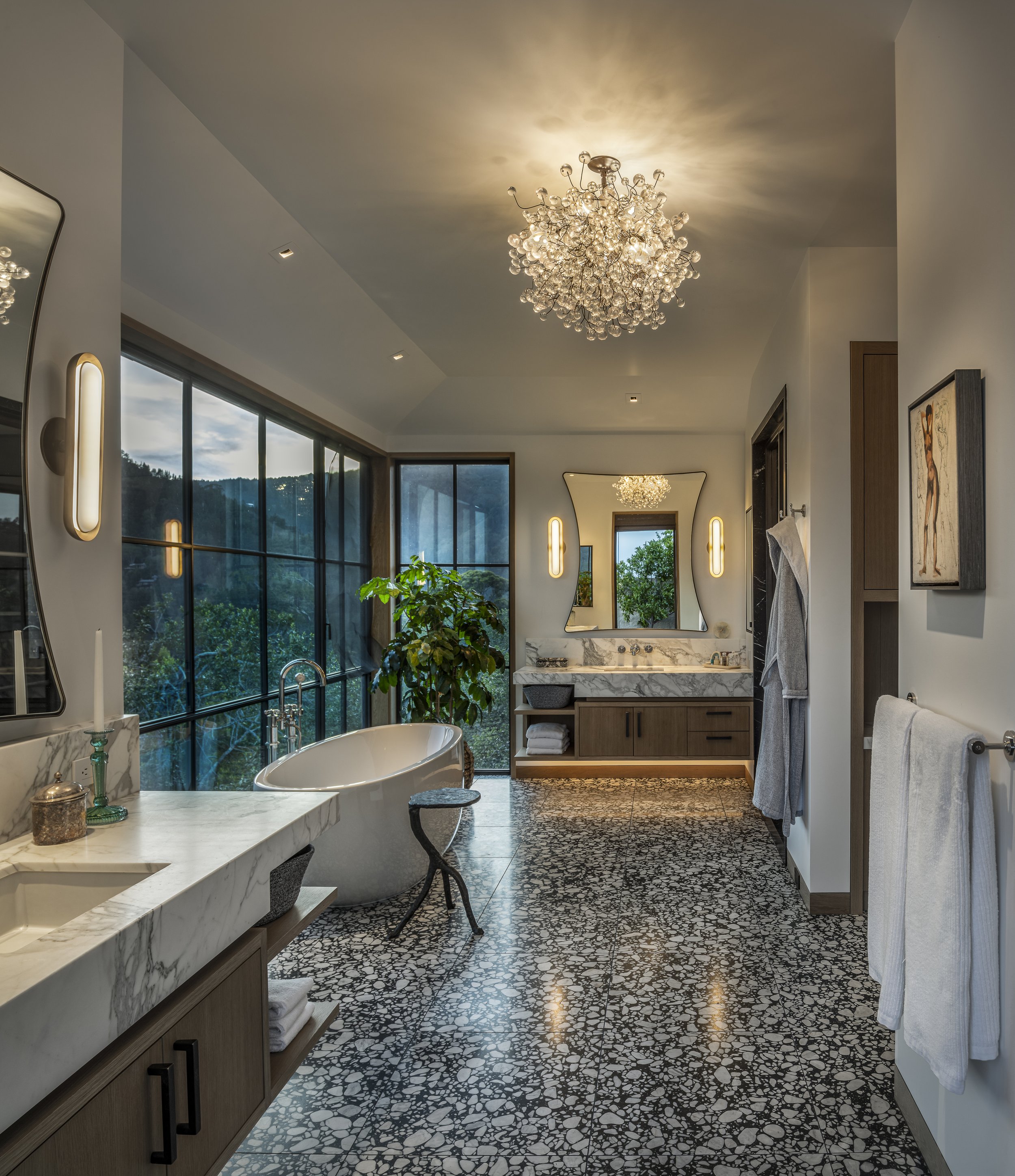 Elegant bathroom with large windows, stone floor, marble countertop, and chandelier lighting.