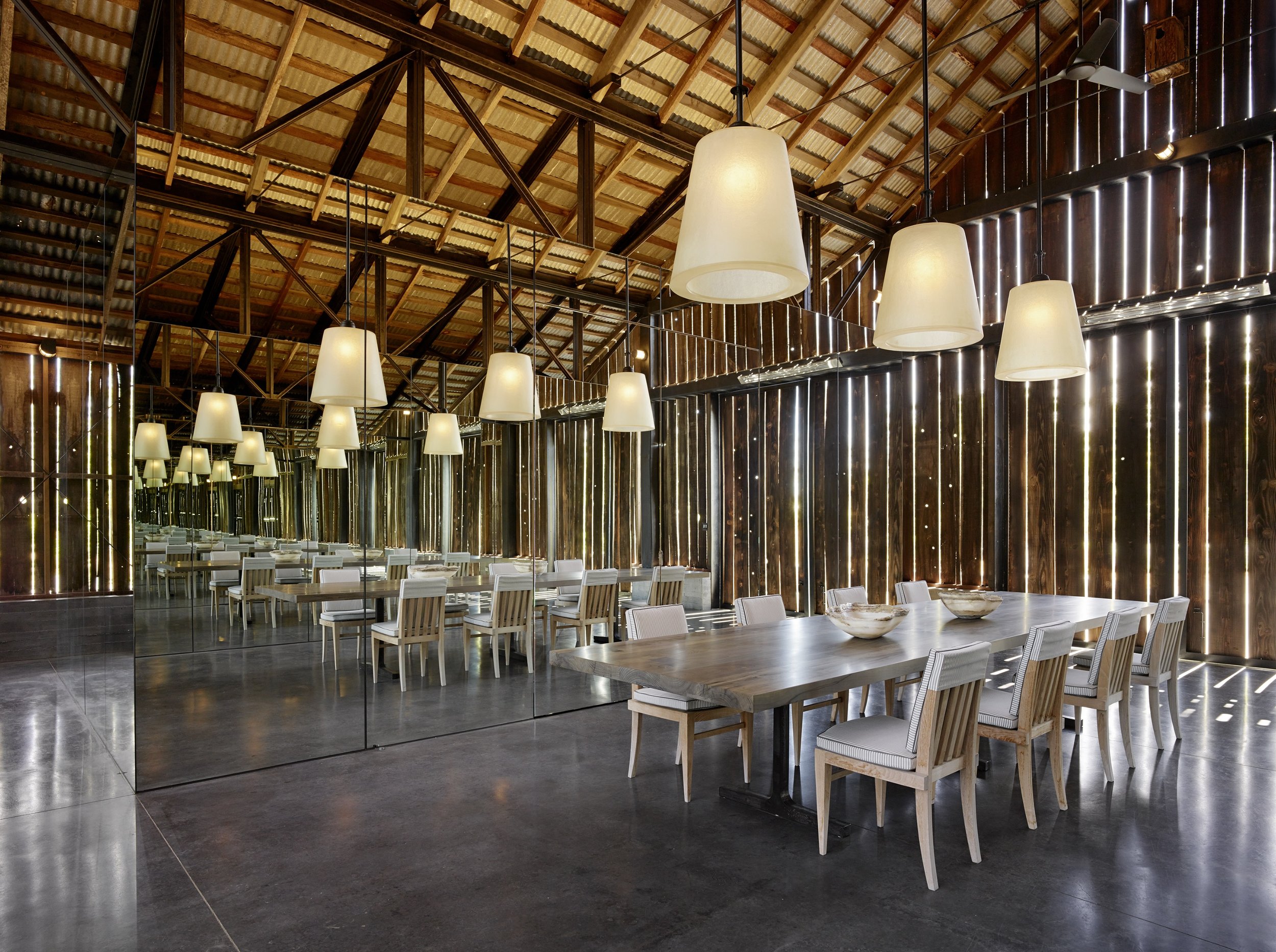 A spacious indoor dining area with high wooden ceiling beams, hanging pendant lights, and a reflective wall with vertical slats. There are white chairs around wooden tables, and the floor appears to be polished concrete.
