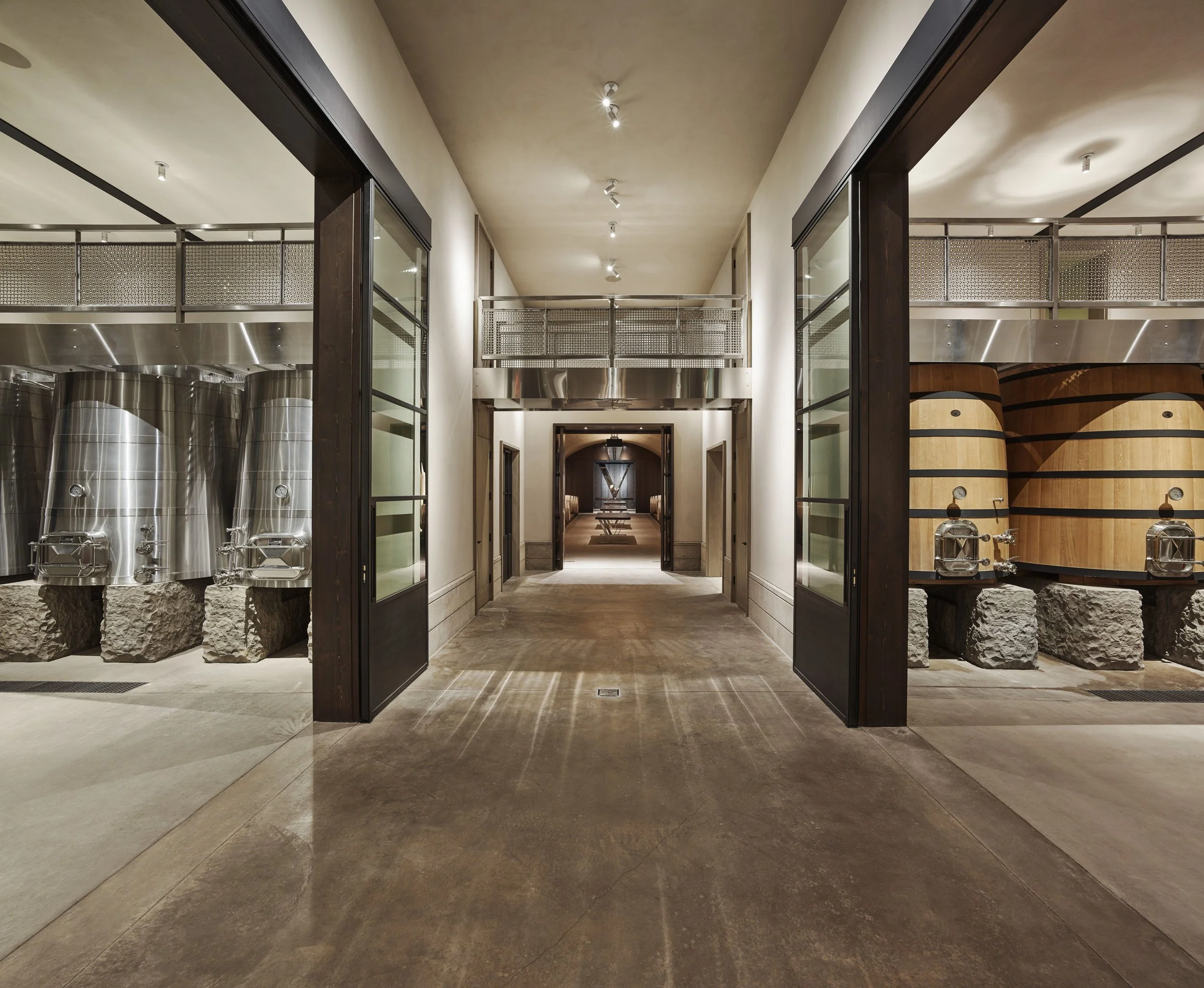 Interior view of a winery, featuring stainless steel fermentation tanks on the left, wooden barrels on the right, and a hallway with a door at the end.