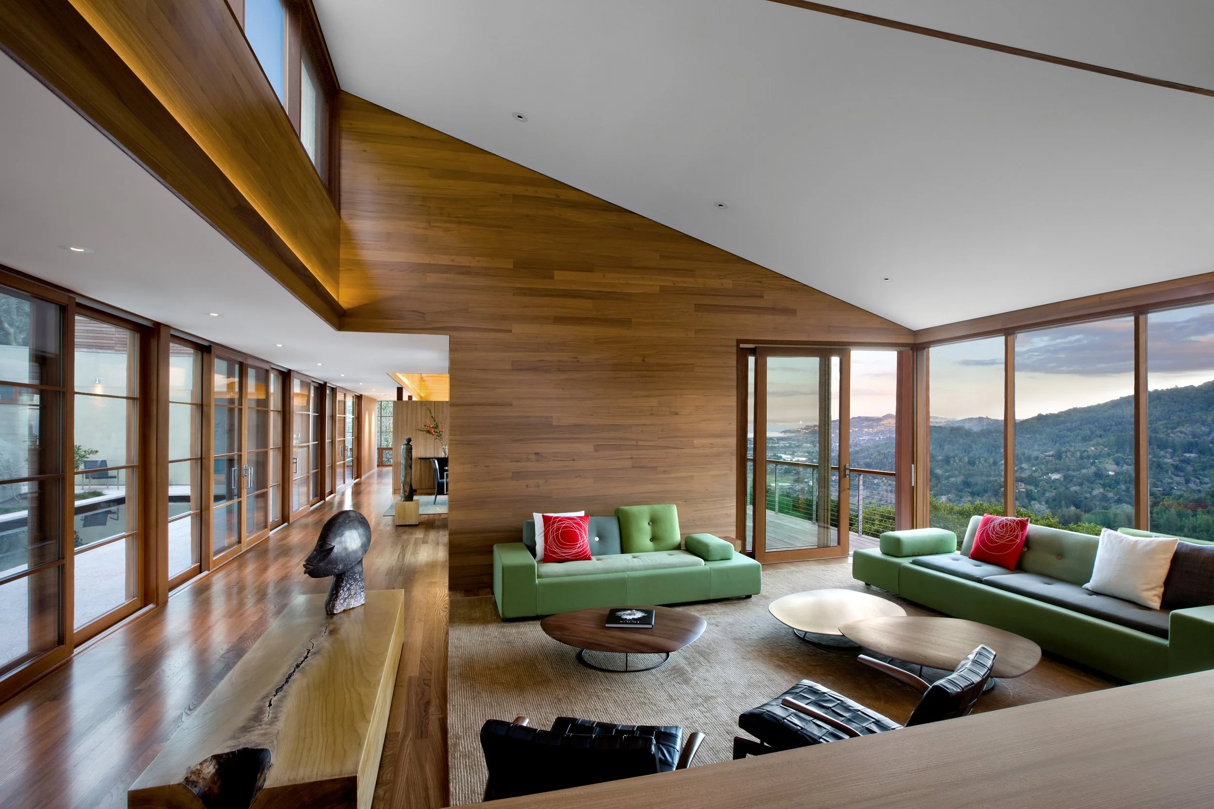 Modern living room with large windows, wooden ceiling and floor, green sofas with red pillows, and scenic mountain view outside.