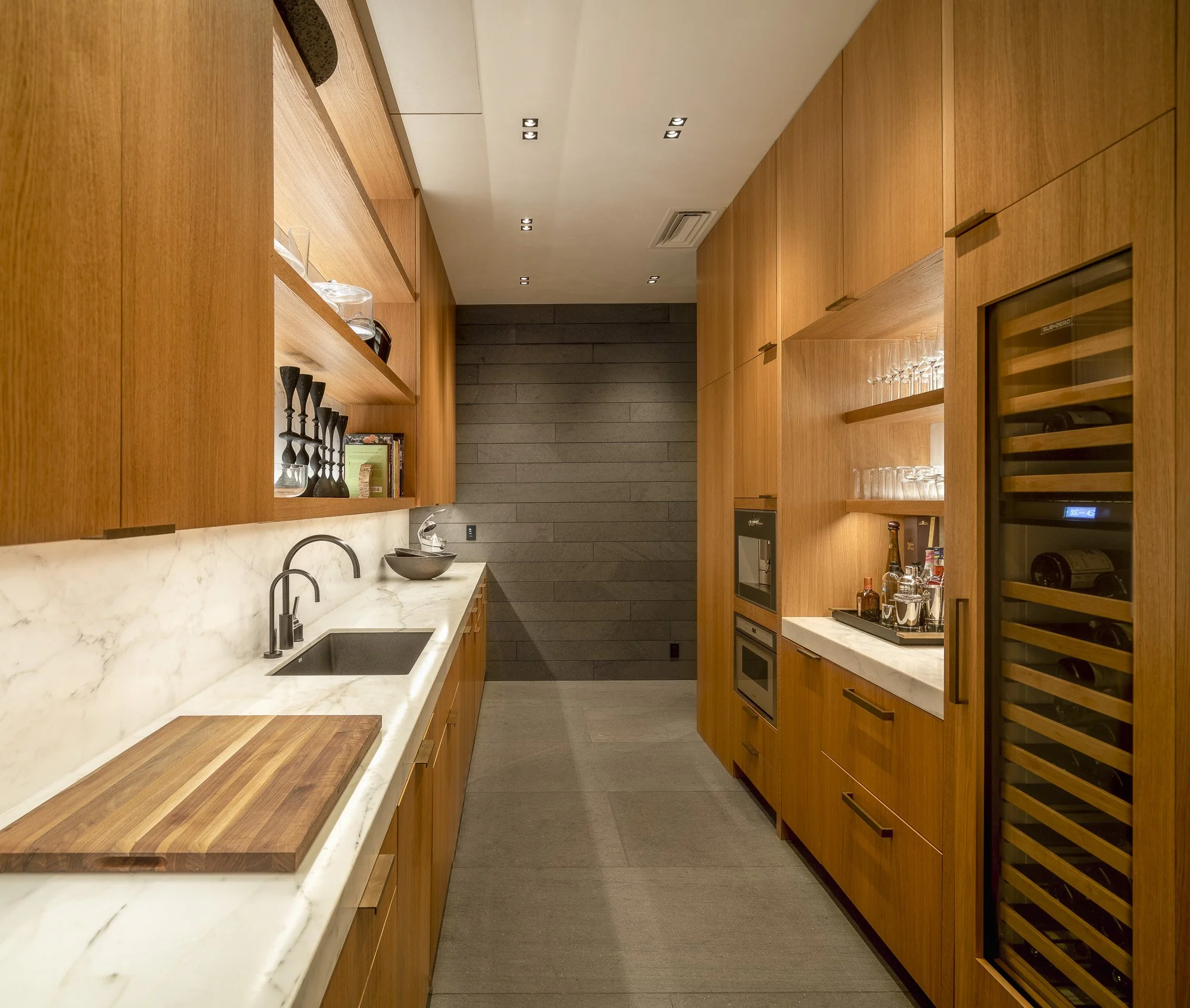 Modern kitchen with wooden cabinets, white marble countertops, black sink, wine fridge, glassware, and a gray tiled wall at the end.