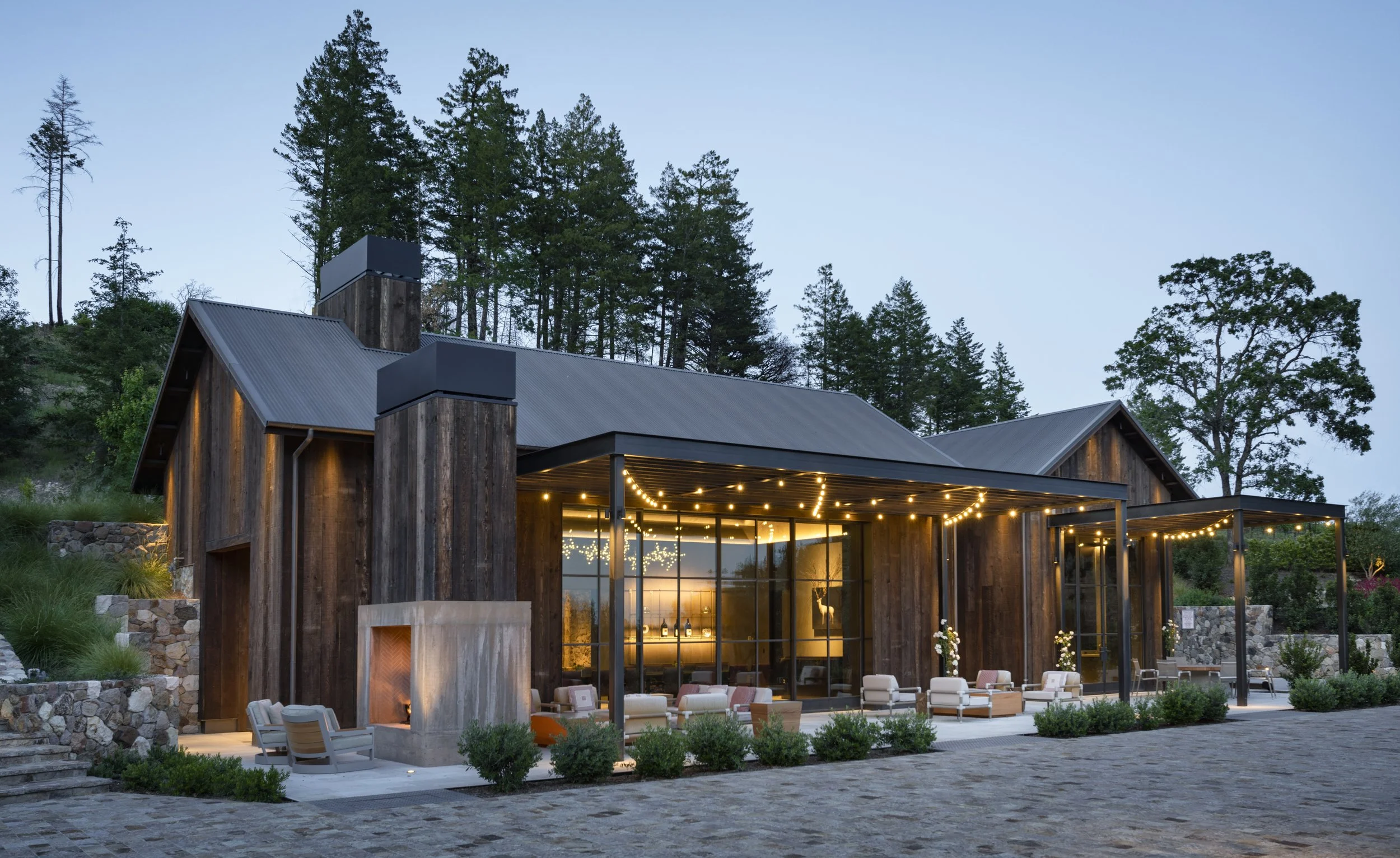 A modern house with large glass windows and a metal roof, surrounded by trees and outdoor seating with string lights at dusk.