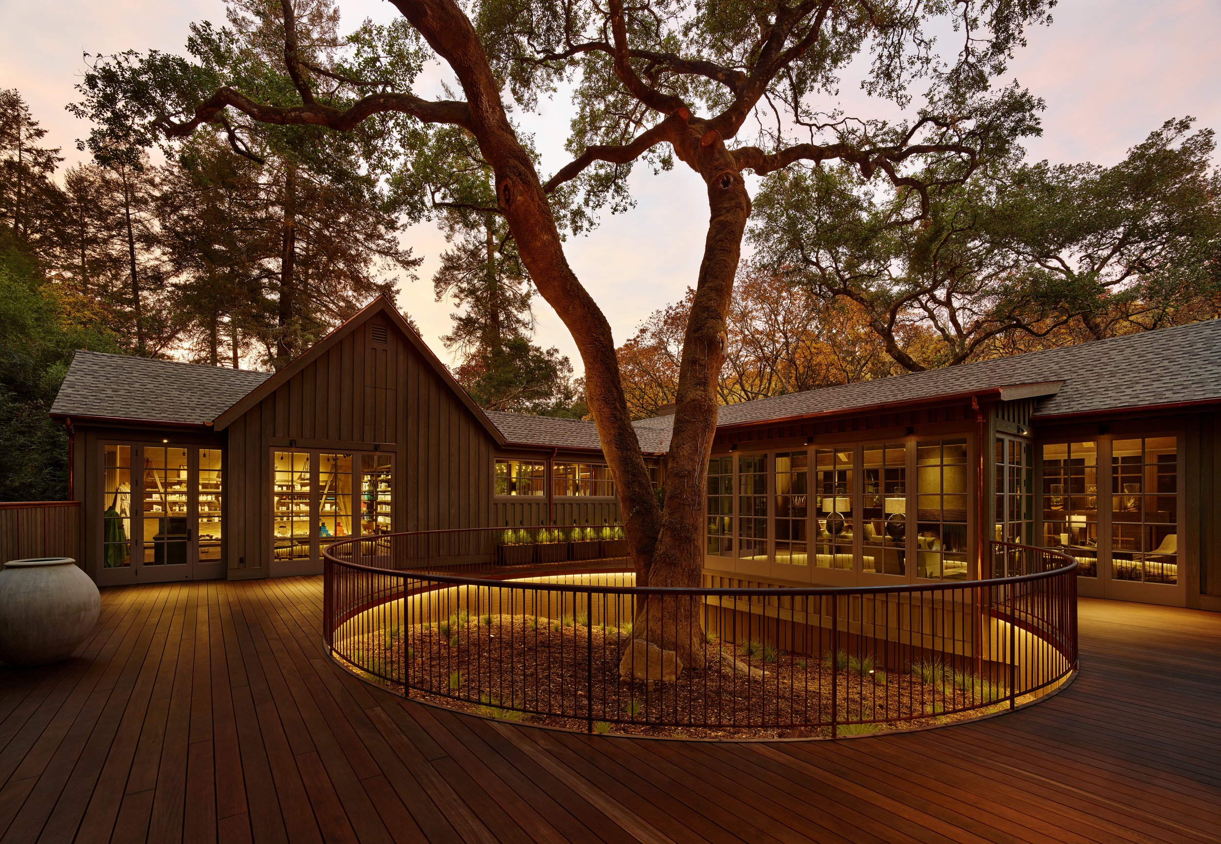 A wooden house with large glass windows at sunset, surrounded by trees, a deck with a railing, and a garden area with a tree and rocks.