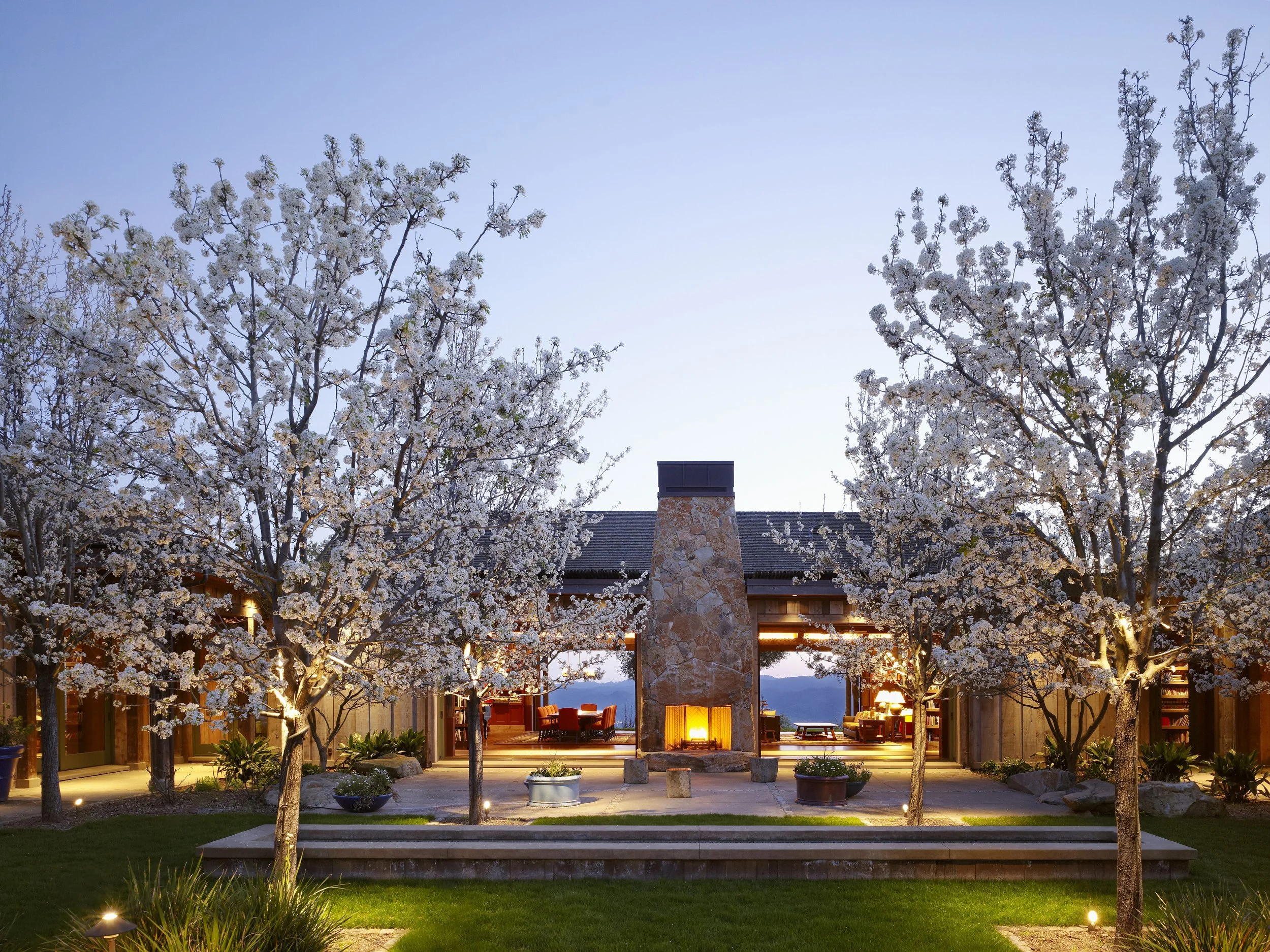 Night view of a house with a stone fireplace, large windows, surrounded by blossoming trees and outdoor lighting