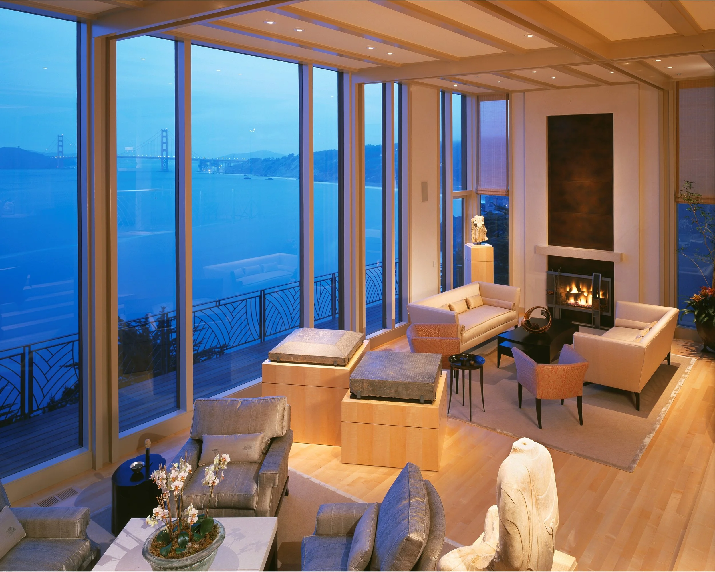 Living room with large floor-to-ceiling windows showing a waterfront view of the Golden Gate Bridge and hills at twilight, furnished with beige sofas, an armchair, a television, a fireplace, and plants.