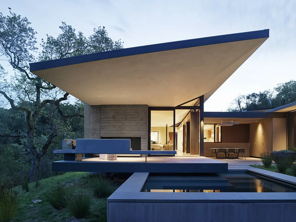 Modern house with large overhanging roof, glass doors, outdoor seating, and a swimming pool in a natural setting during dusk.
