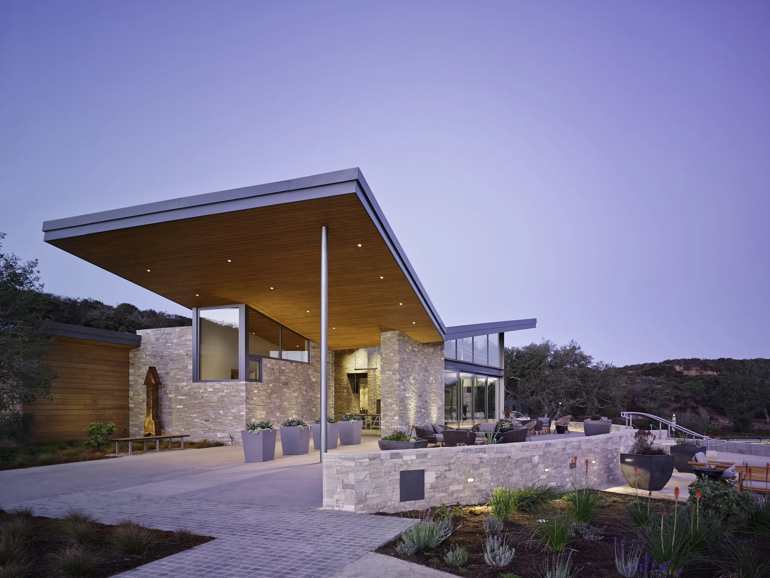 Modern house with large glass windows and wooden and stone exterior, outdoor patio area with seating, planters, and yard with plants, set against a backdrop of hills and trees during dusk.