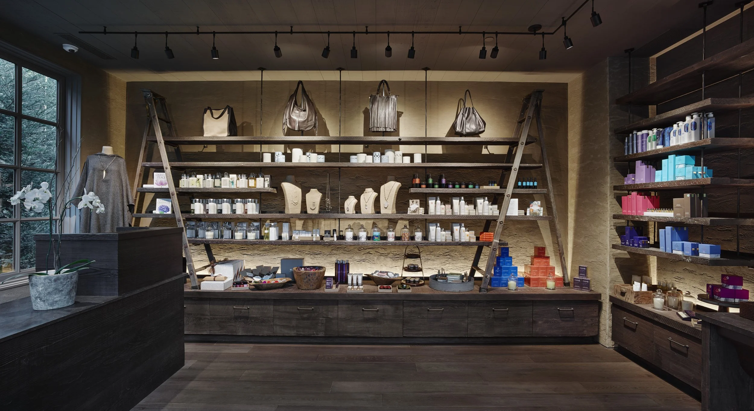 Interior of a retail store with wooden shelves displaying handbags, jewelry, candles, and skincare products. A large window on the left lets in natural light, and there is a plant with white flowers near the window.