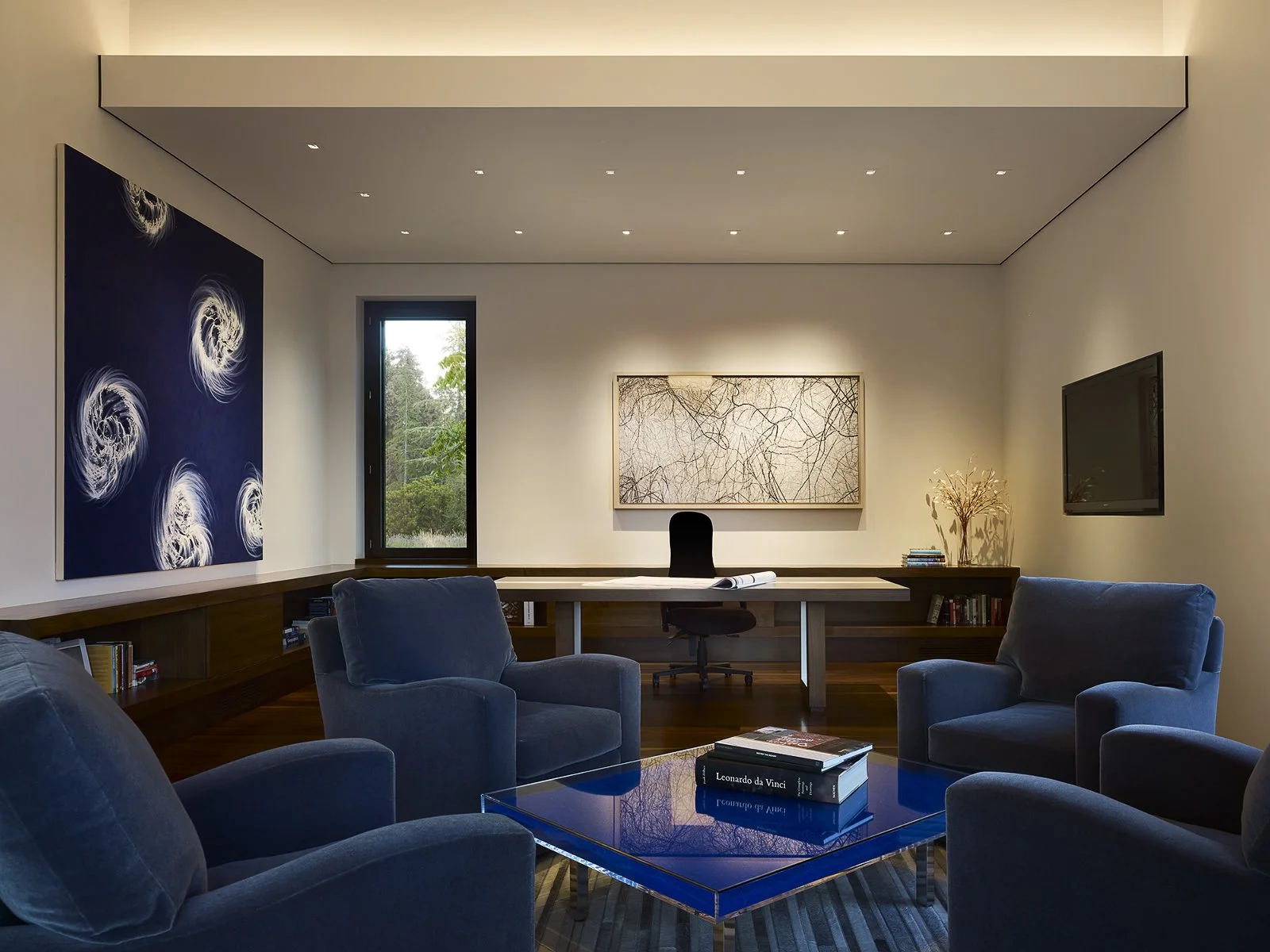 Modern living room with blue armchairs, a coffee table with books, wall art, desk with a black chair, and a large window showcasing greenery outside.