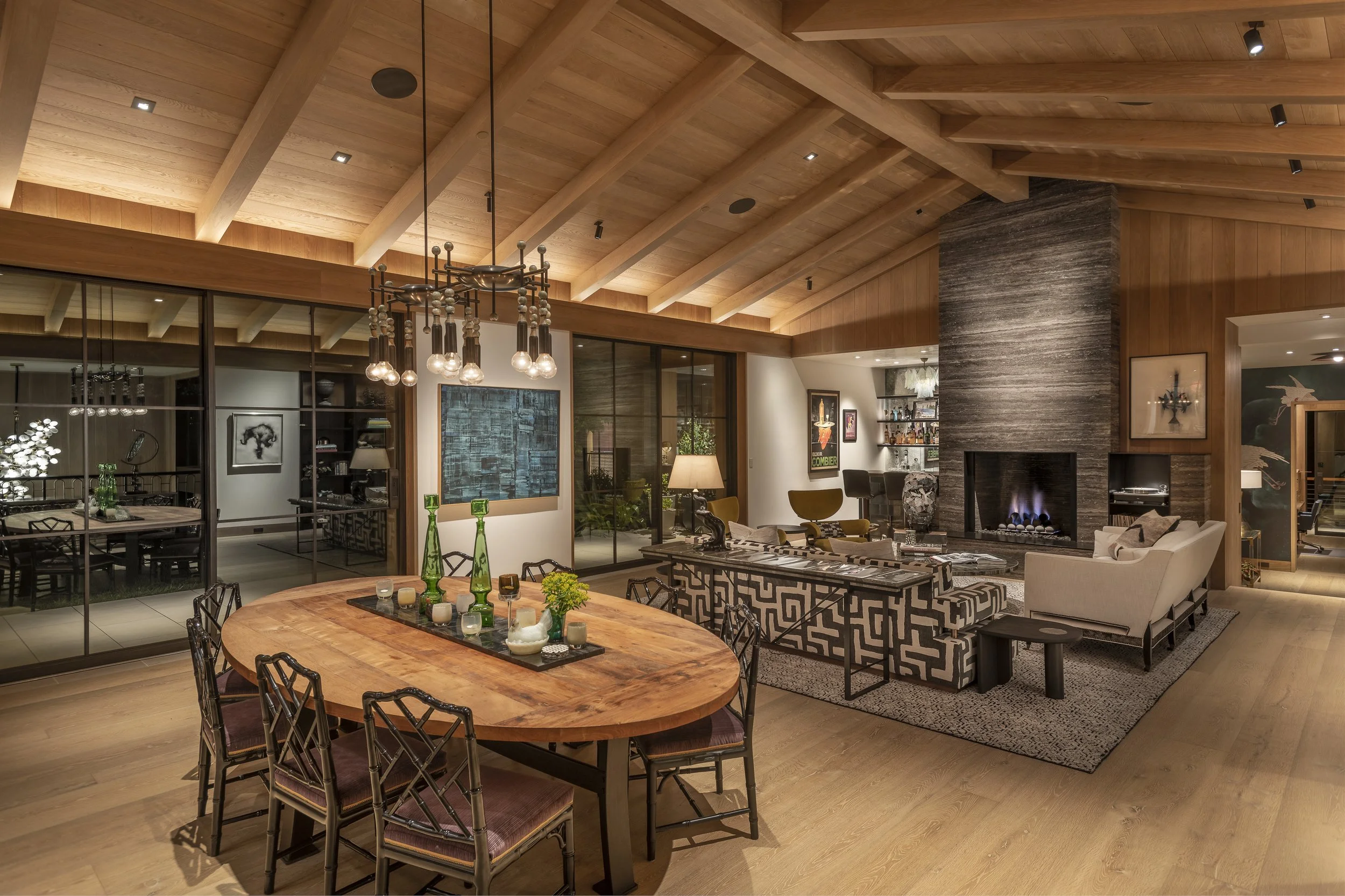 Modern open-concept living room and dining area with wooden ceiling beams, a stone fireplace, glass sliding doors, and contemporary furniture.