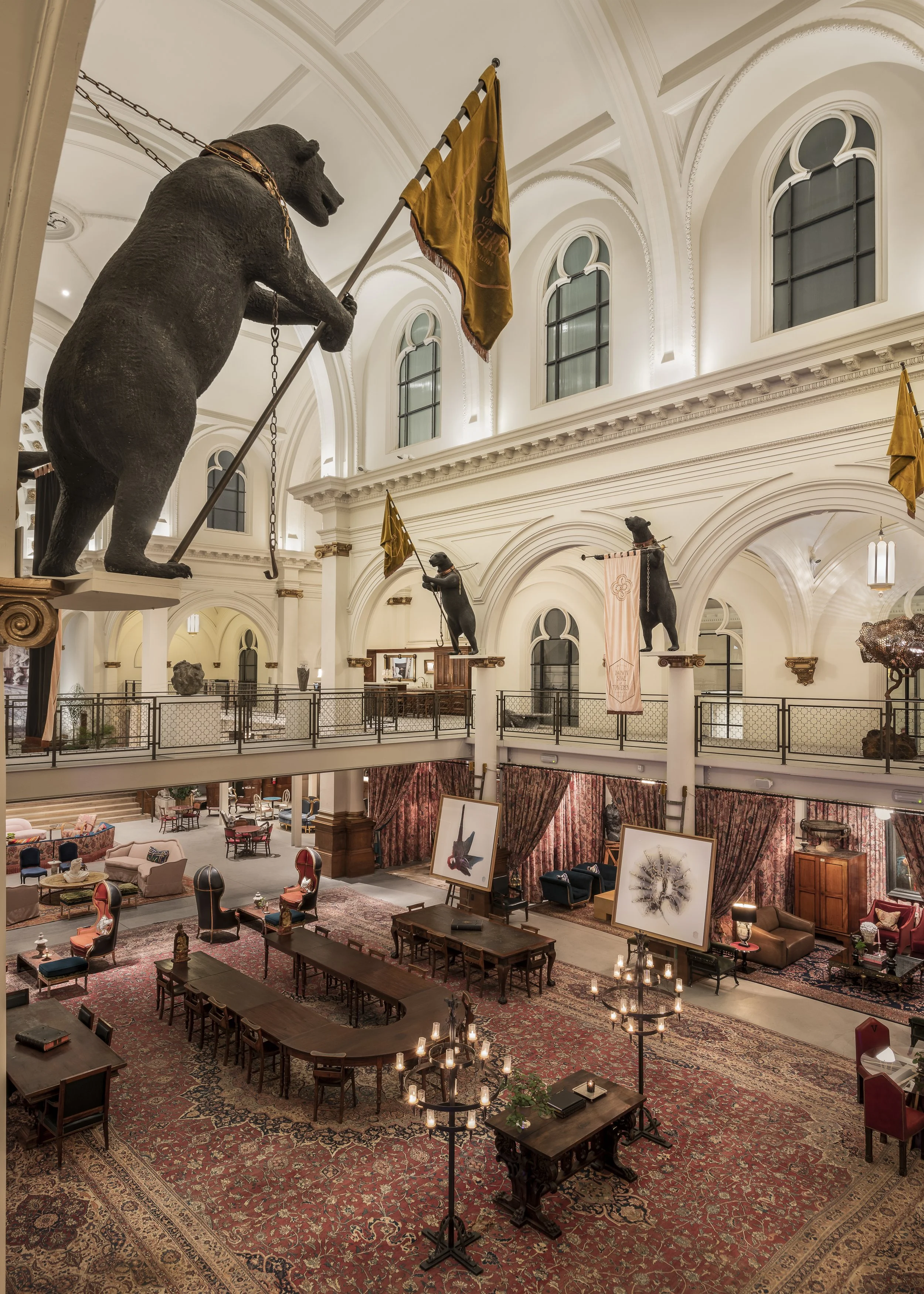 Interior view of a grand, elegant room with high arched windows, displaying large black bear statues holding flags, and various vintage furniture, paintings, and decorative items in a classical style.