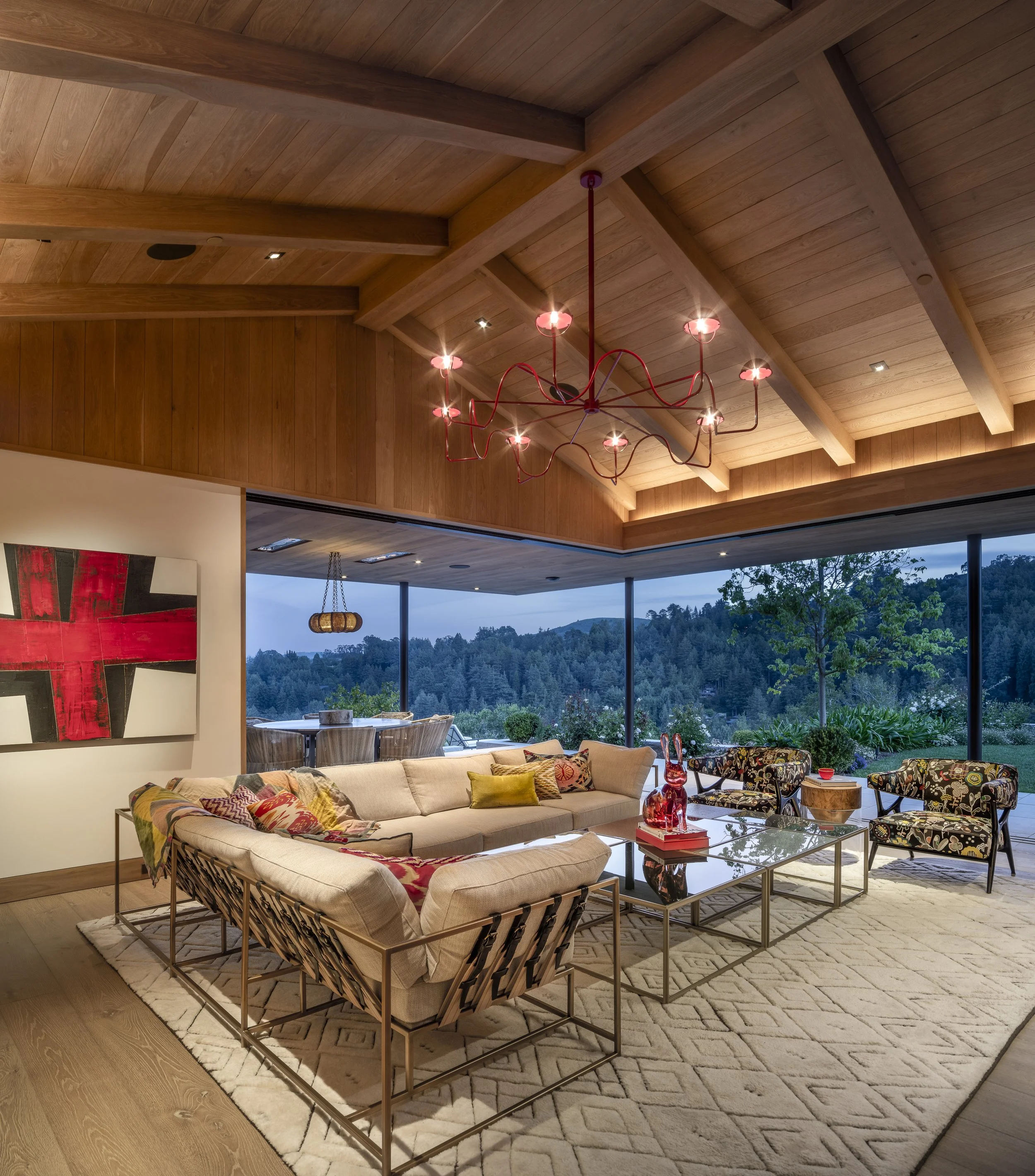 Living room with large cream sofa, colorful pillows, glass coffee table with red decorative items, patterned armchairs, area rug, wooden ceiling with beams, red modern chandelier, large windows showing outdoor landscape.