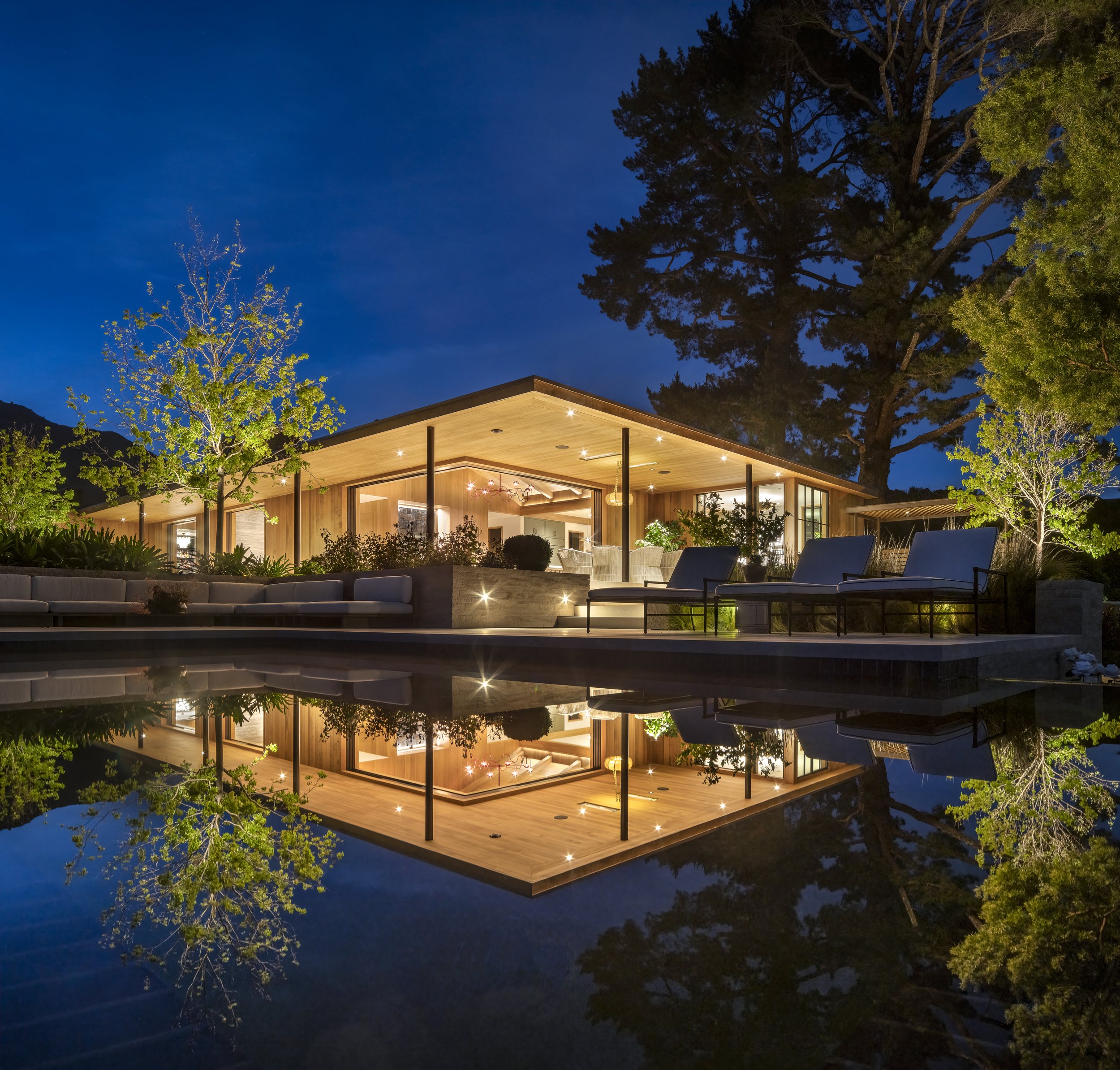 Nighttime view of a modern house with large windows and outdoor lighting, surrounded by trees, and reflected in a pool of water.