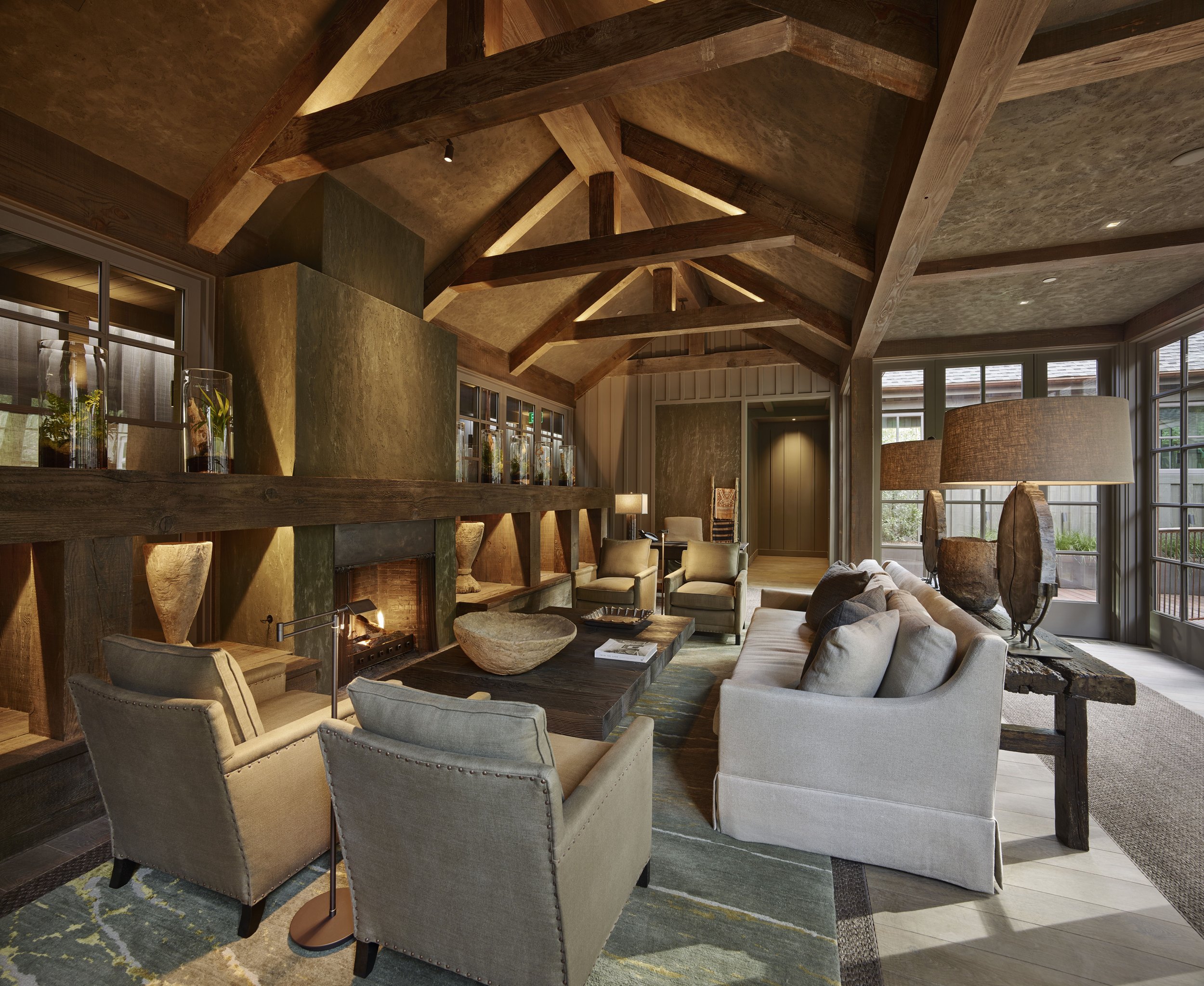 Interior of a living room with a rustic wooden vaulted ceiling, large windows, beige upholstered chairs, and a fireplace with decorative glass vases on the mantle.