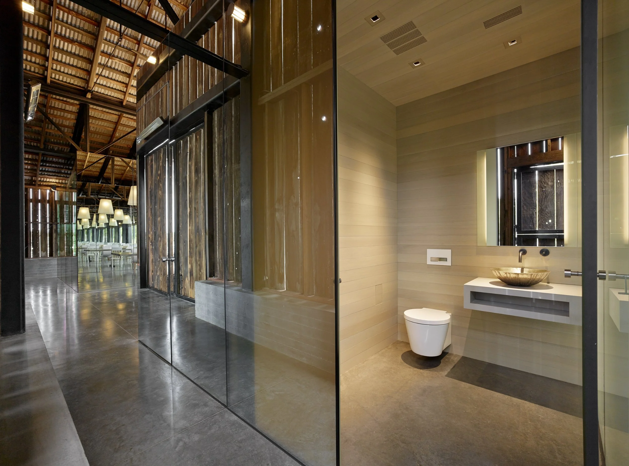 Interior of a modern bathroom with a wall-mounted toilet, a mirror, and a stone sink, separated by a glass wall from a large room with wooden walls and vaulted ceilings, lit by pendant lights and wall sconces.