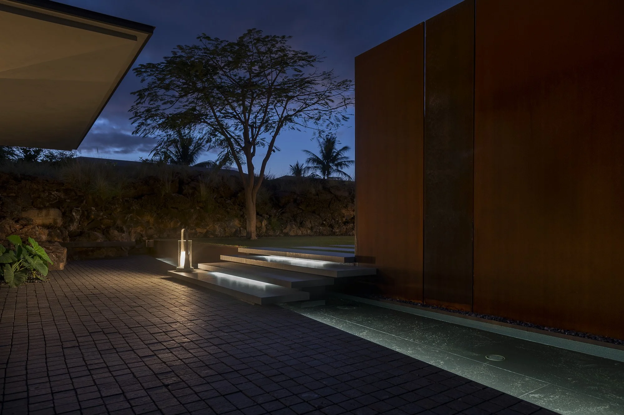 A modern outdoor yard at dusk with a tree, a fountain, steps, and a building wall illuminated by subtle lighting.