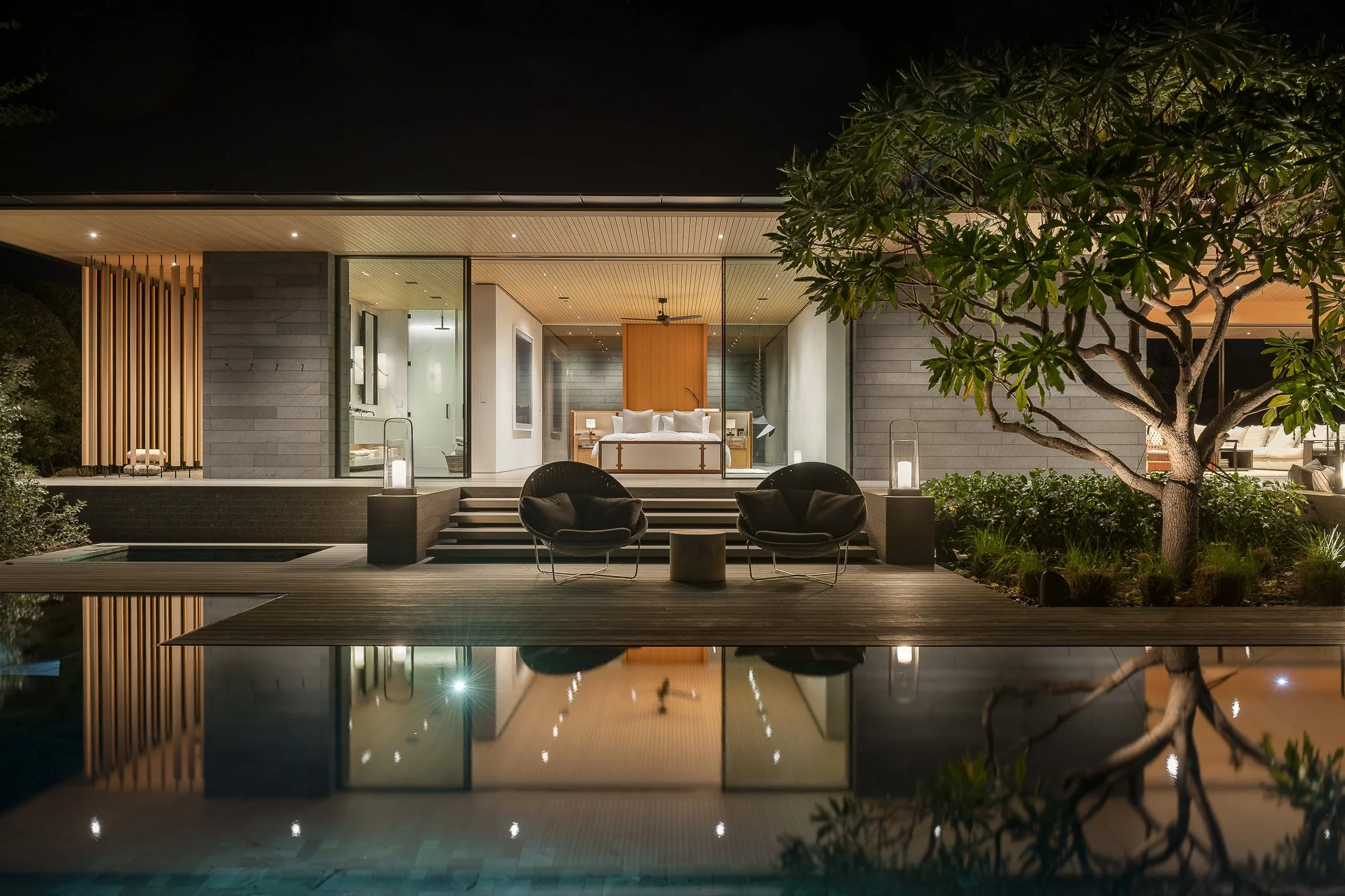 Nighttime view of a modern house with illuminated interior, a swimming pool reflecting the house, outdoor seating, a large tree on the right, and landscaped garden.