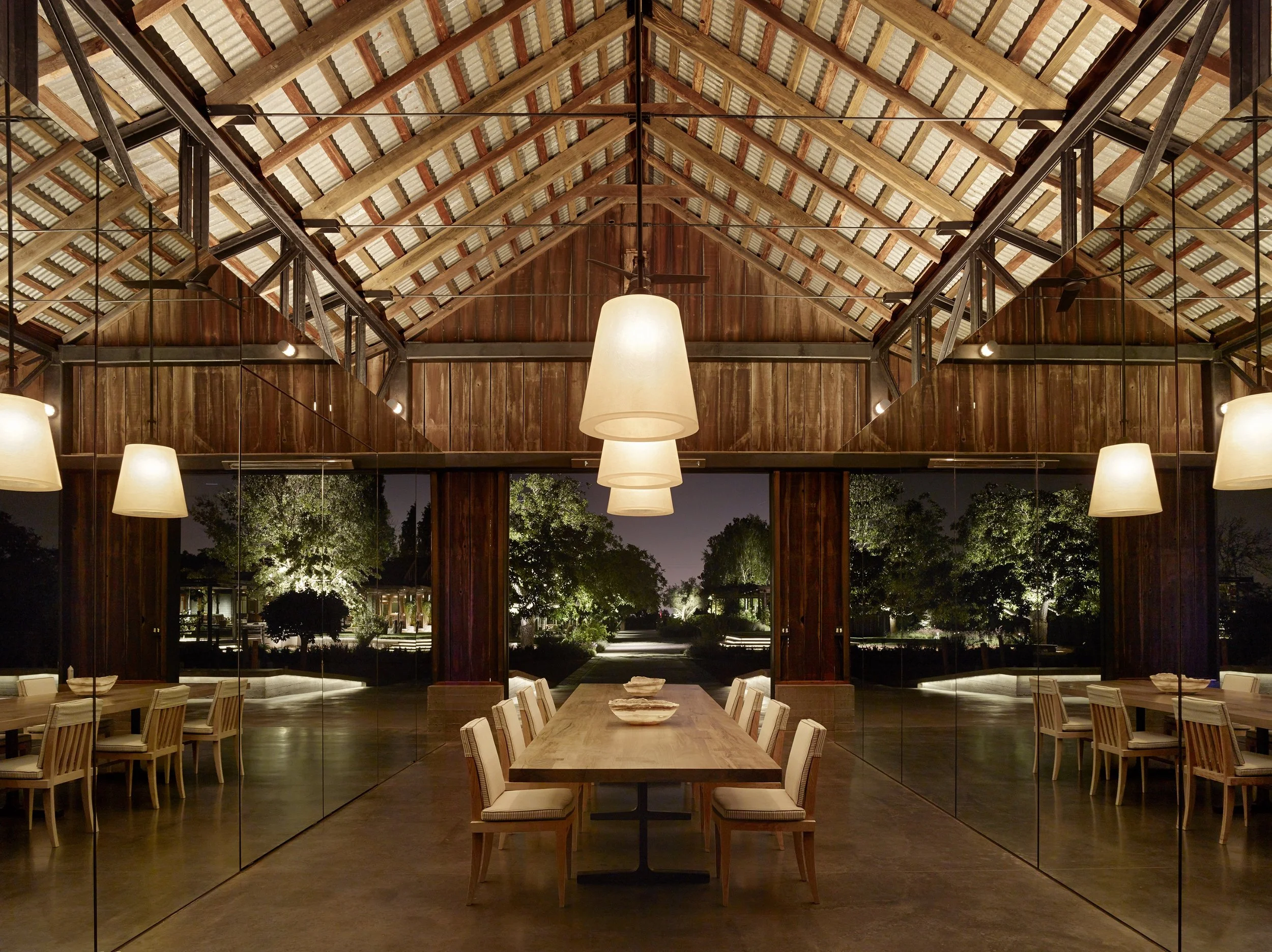 Interior view of a wooden dining area with a vaulted ceiling, hanging pendant lights, and a long wooden table with chairs, overlooking an outdoor night scene with trees.