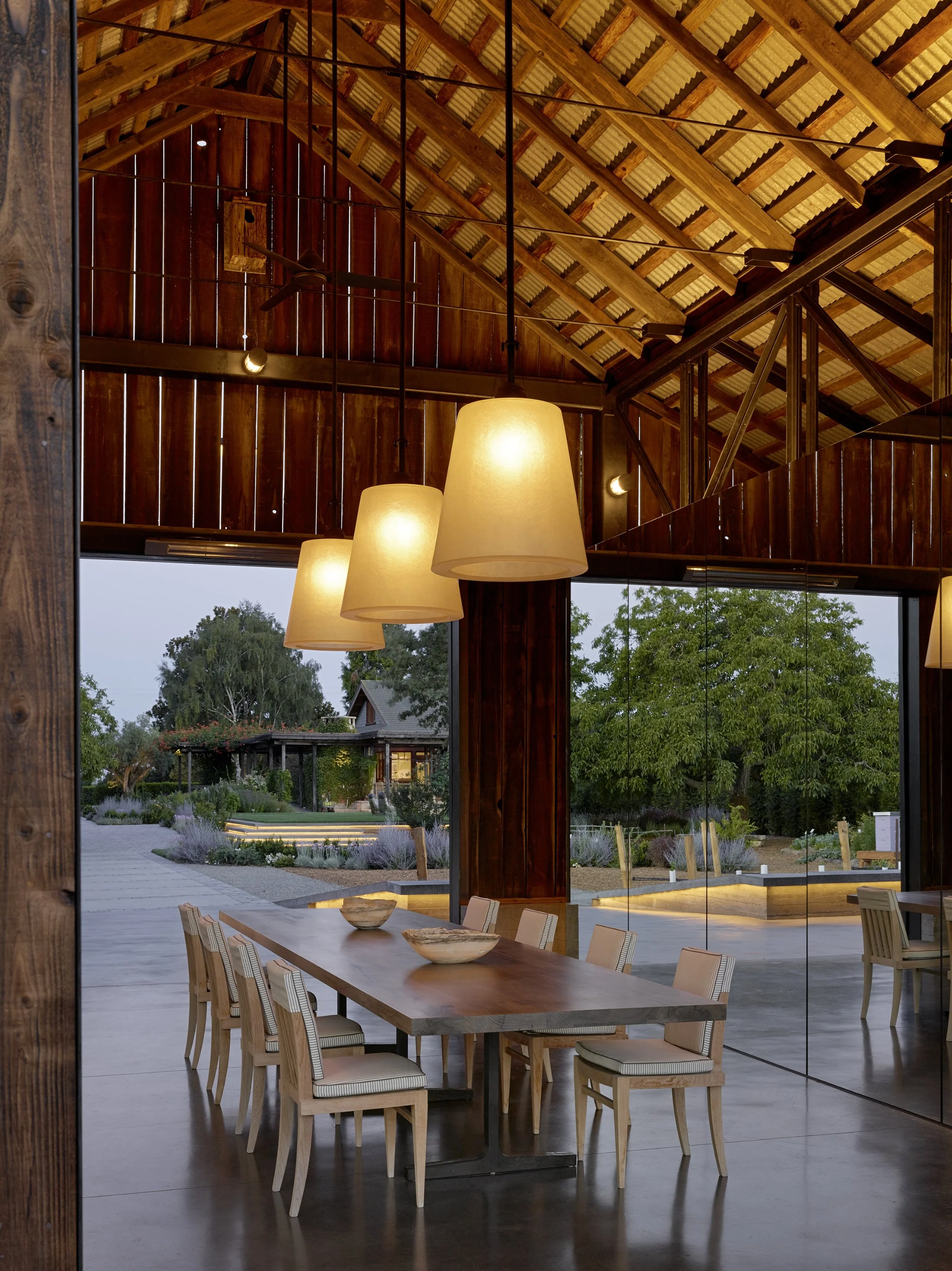 Interior view of a modern wooden dining area with a table, chairs, hanging lamps, and a glass wall showing an outdoor garden with trees and plants.