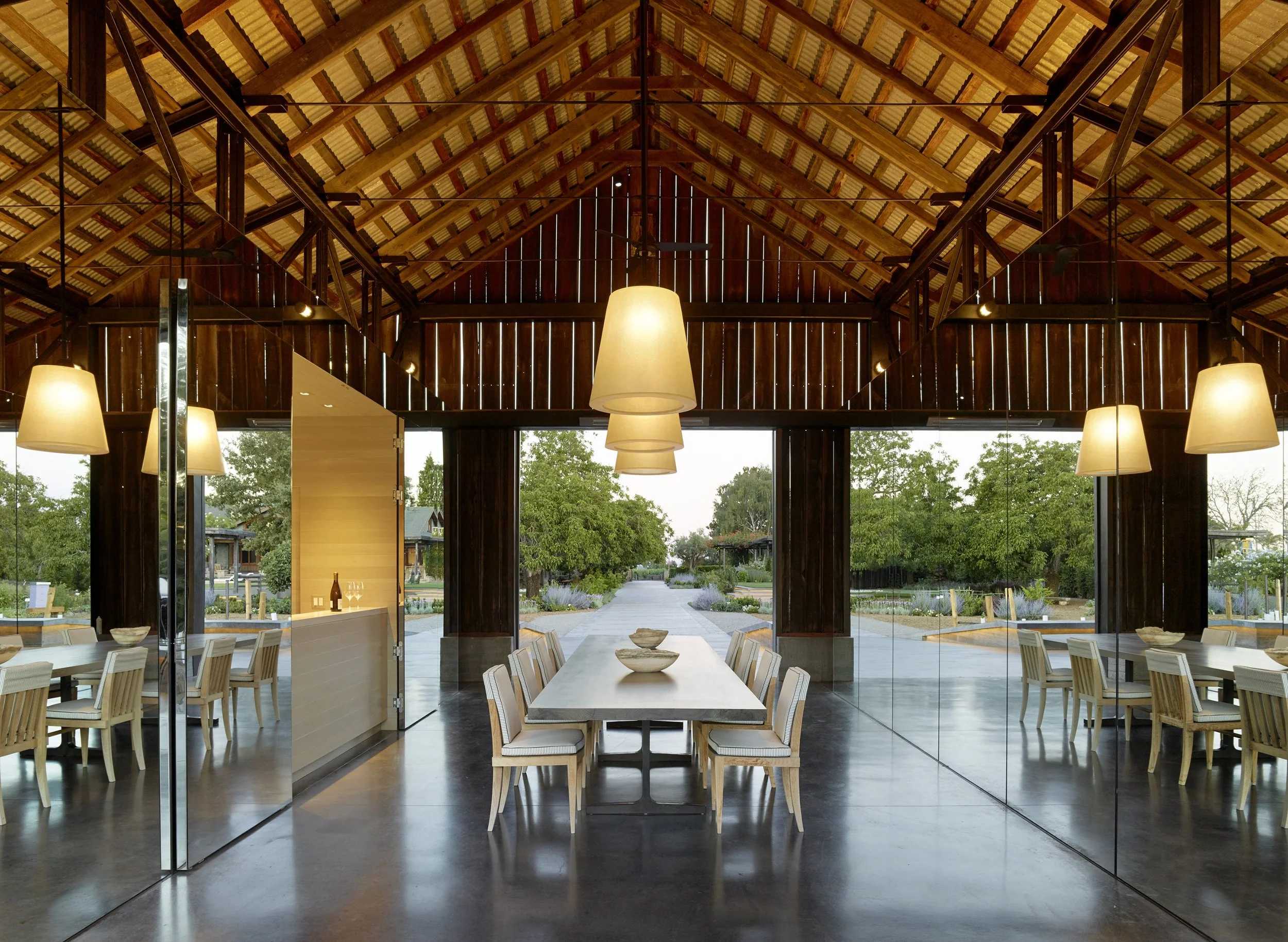 Indoor dining area with wooden ceiling and large glass windows showing outdoor greenery, modern pendant lights hanging above a long dining table surrounded by wooden chairs.
