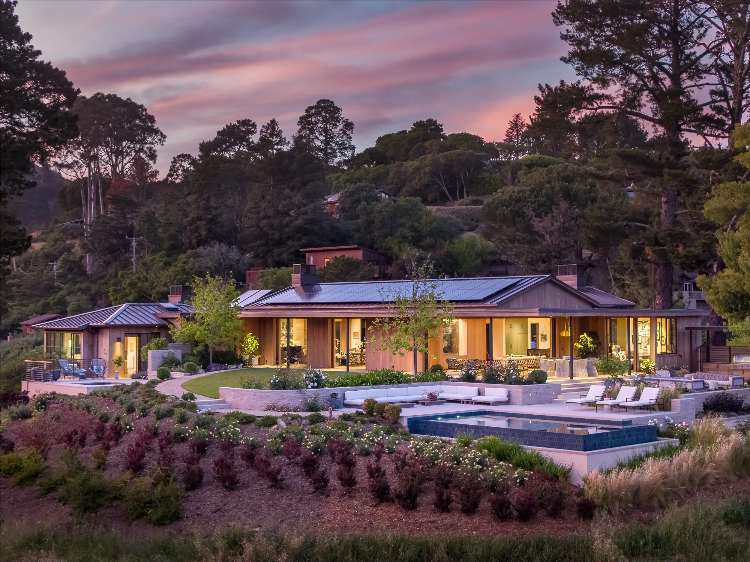 A modern house with a backyard featuring a swimming pool, lounge chairs, and a garden at dusk. The house has large glass windows and is illuminated from inside. In the background, there are trees and a colorful sunset sky.