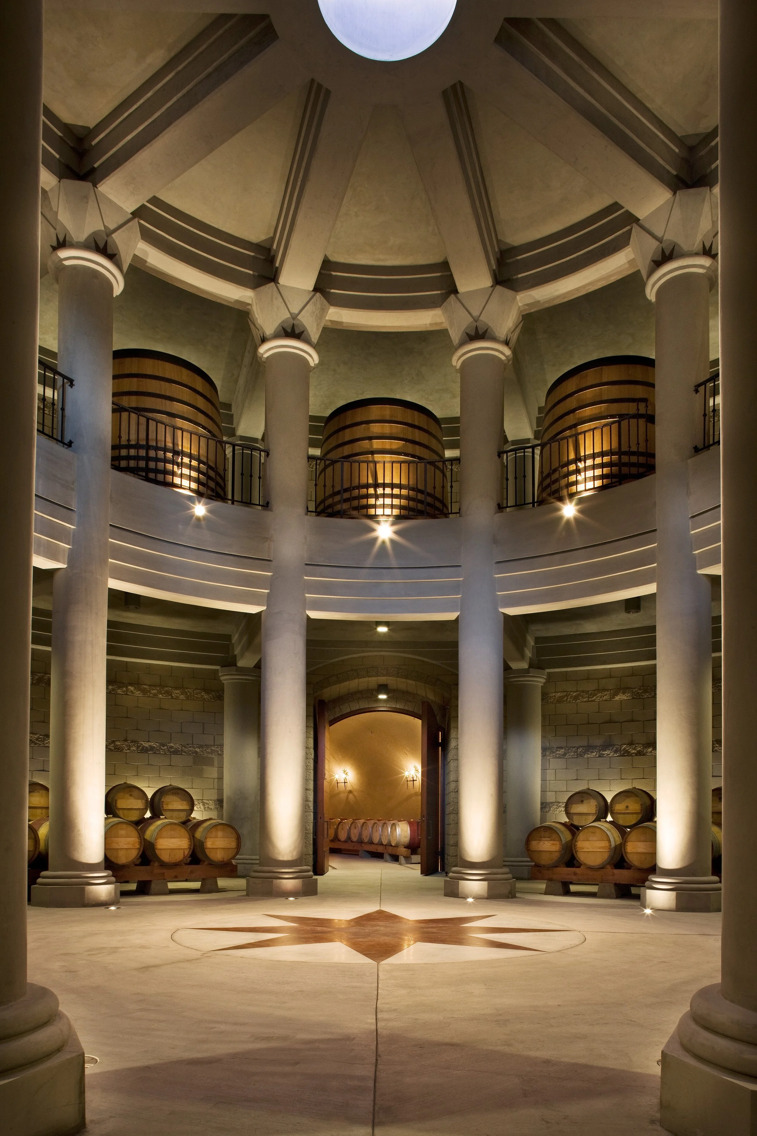 Interior of a winery cellar with large wooden barrels, stone walls, and classical columns.