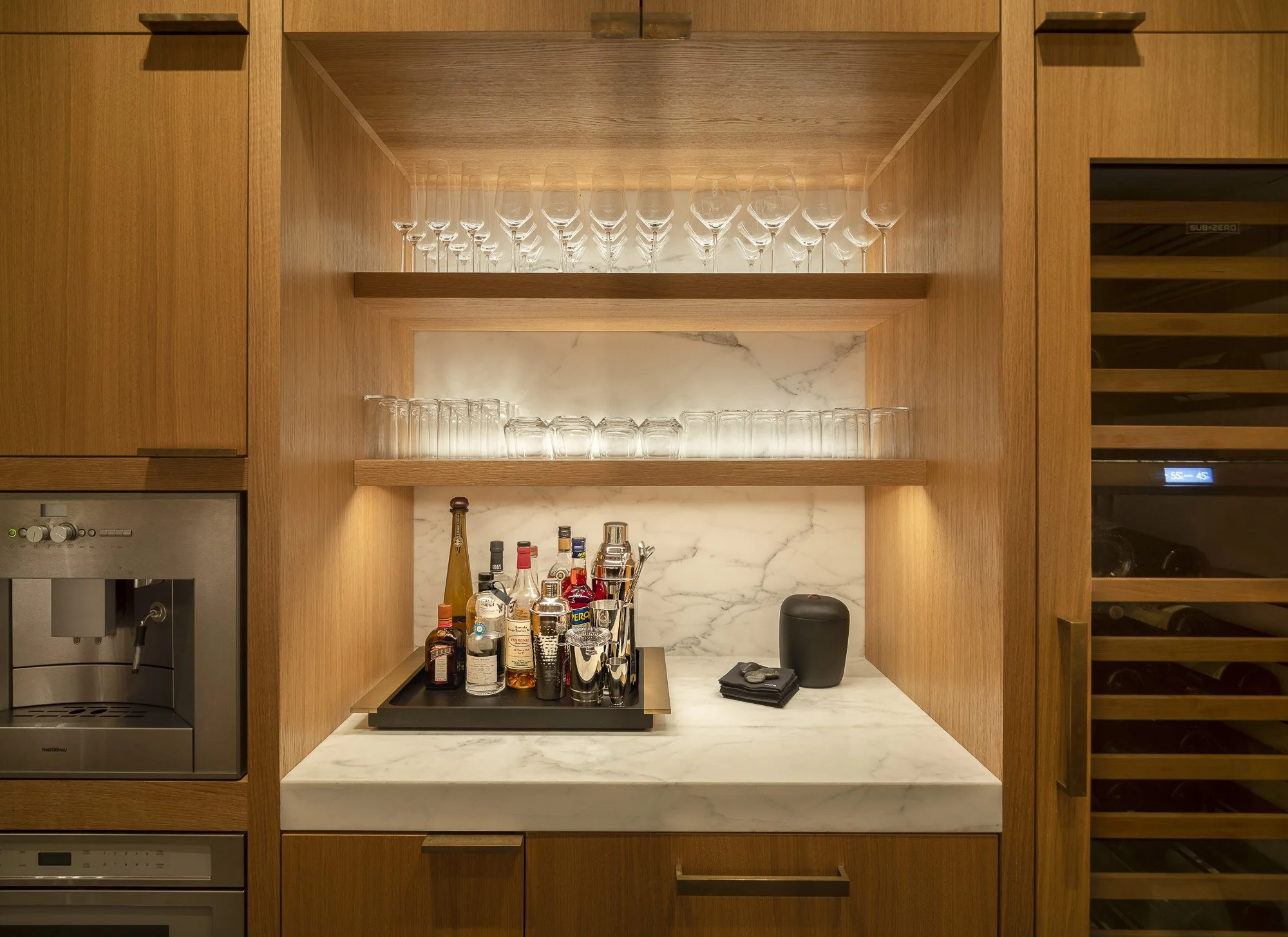 Wooden bar area with shelves holding empty wine glasses and liquor bottles, marble countertop with bar tools and supplies, wine cooler on the right.