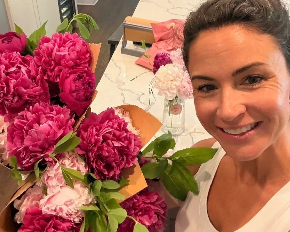 A woman smiling and holding a bouquet of pink peonies in a room with a marble countertop, a glass jar with pink flowers, and various craft supplies.