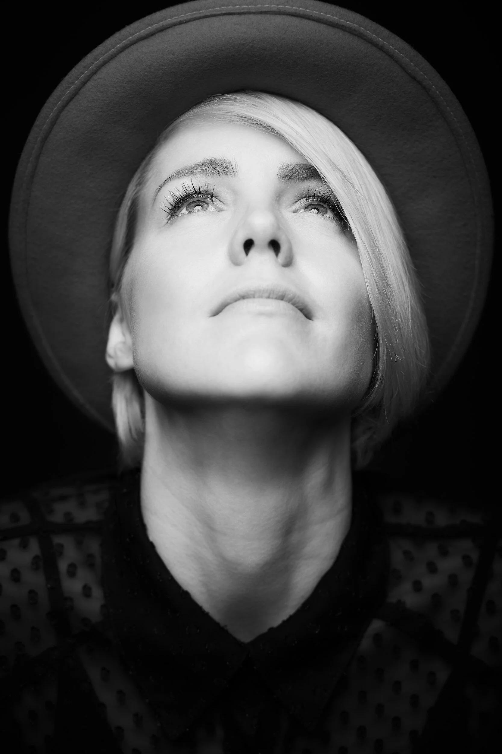 Black and white portrait of a woman with short blonde hair, wearing a wide-brimmed hat and a polka dot blouse, looking upward with a thoughtful expression.