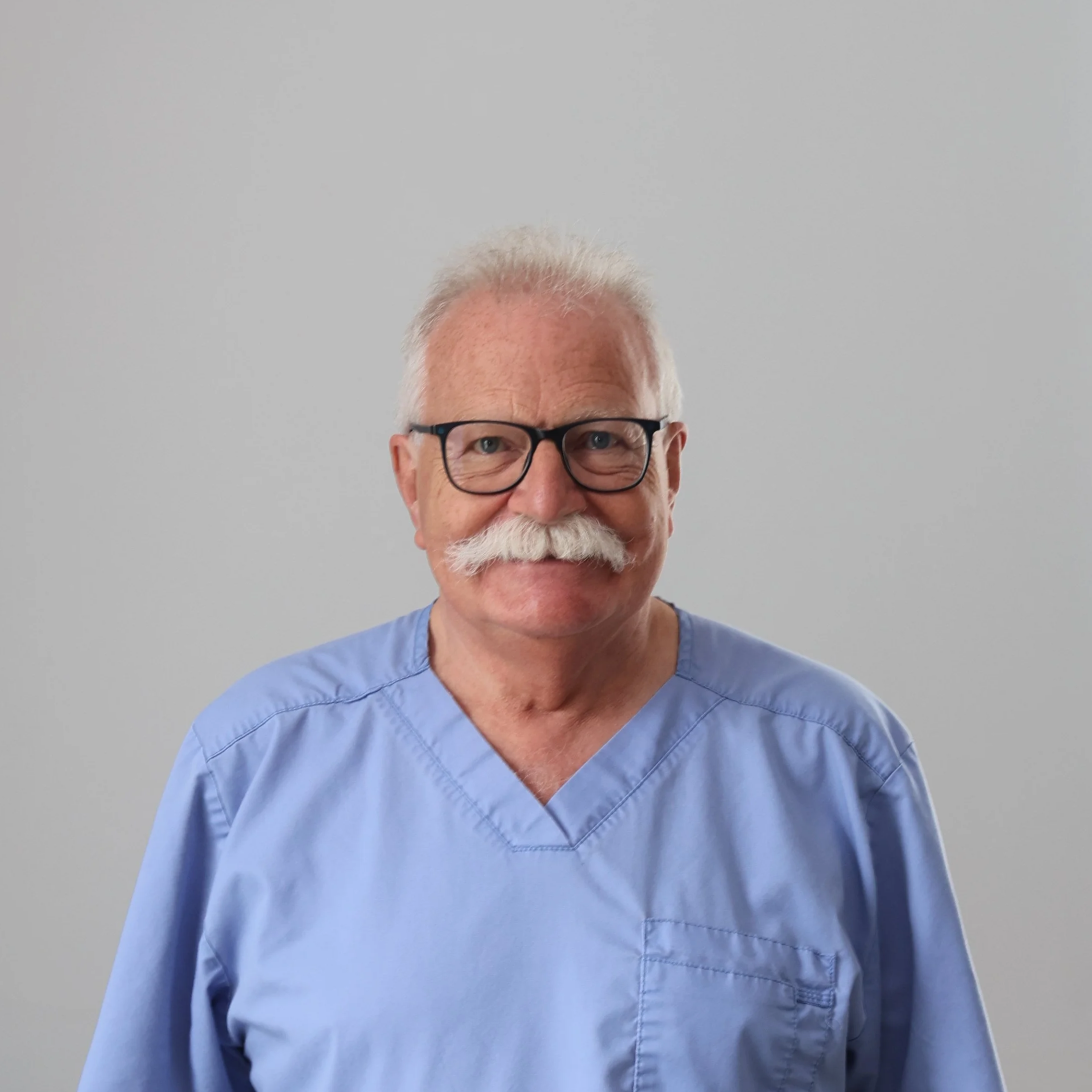 An older man with glasses and a mustache wearing blue medical scrubs in a clinical setting.