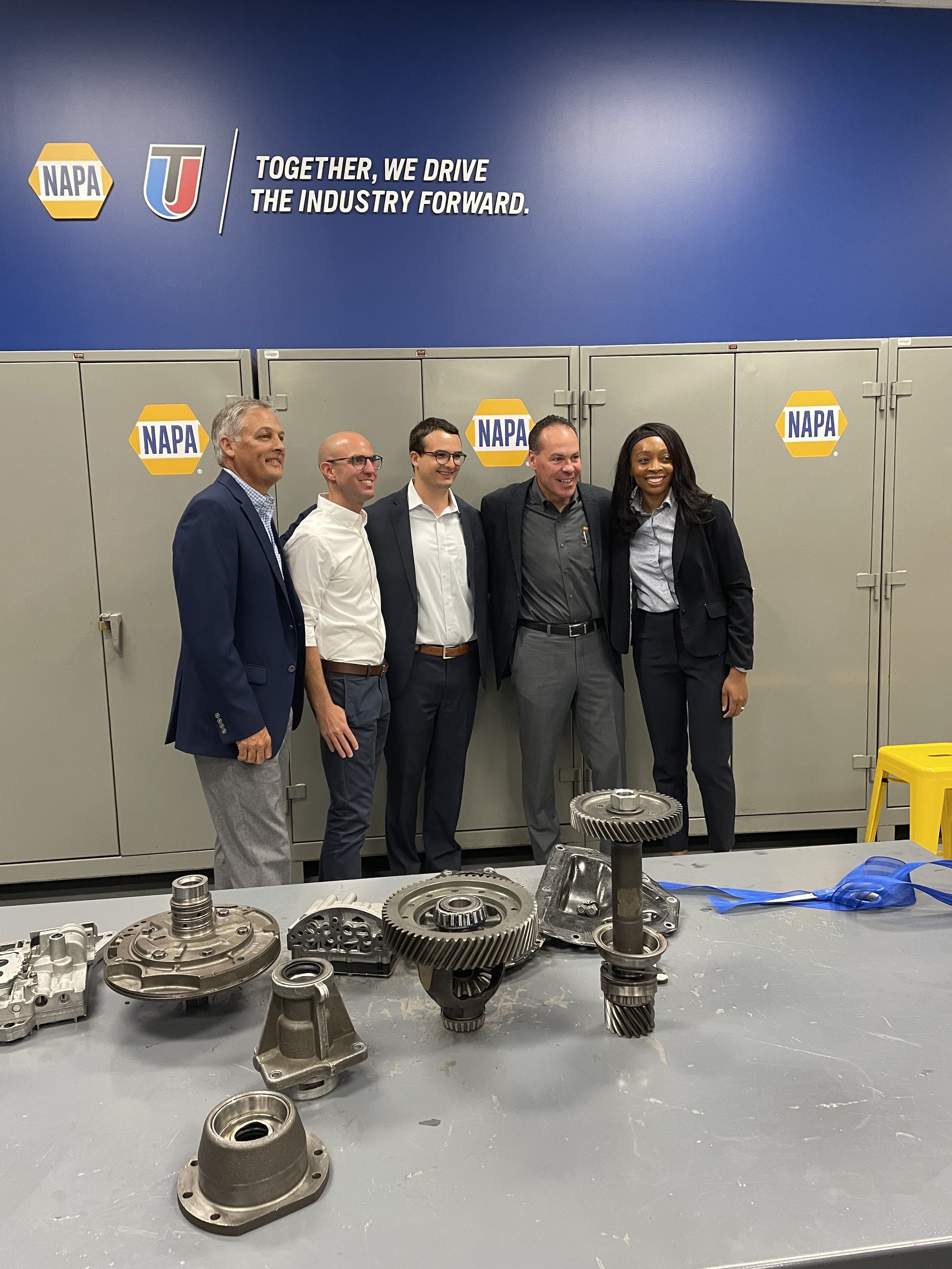 Group of five people in business attire standing in front of storage lockers with NAPA logos at a wheel and automotive parts display.