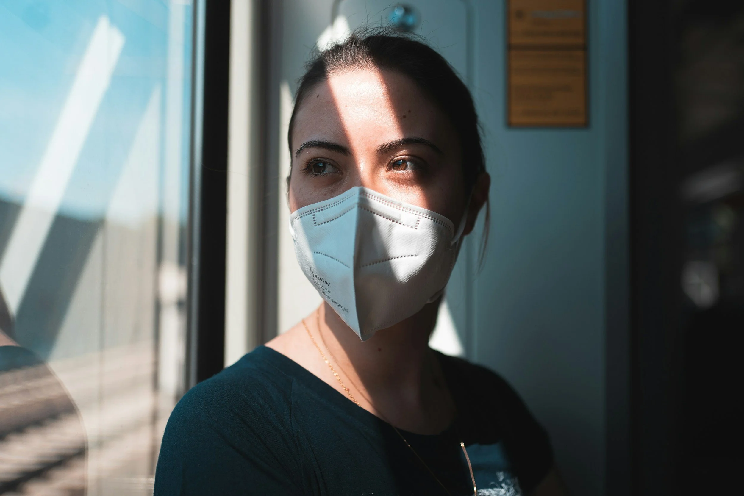 A female nurse wearing a face mask looking out a window with sunlight casting a shadow across her face.