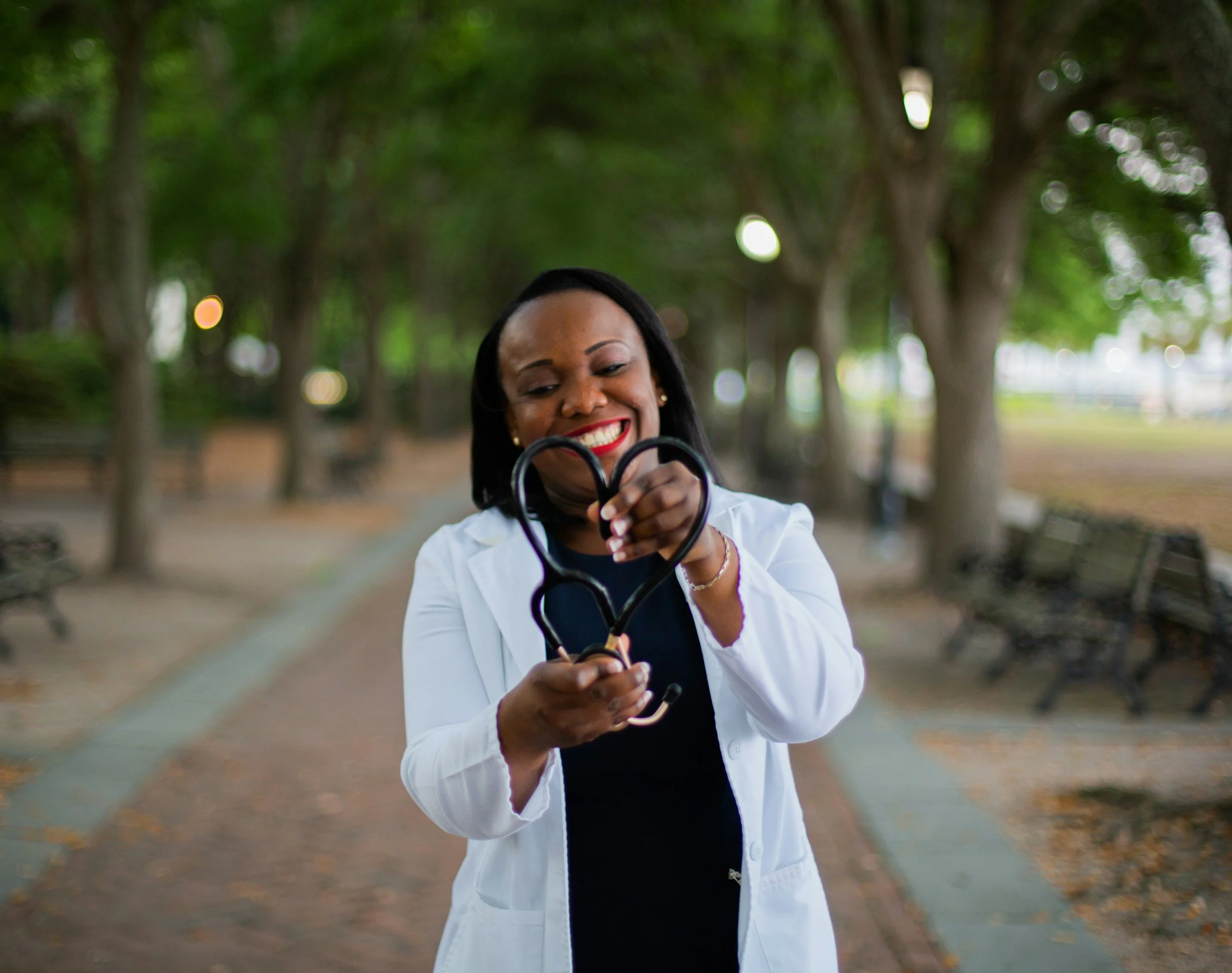 A female doctor in a white coat smiling and holding a stethoscope shaped like a heart in a park with trees and benches.
