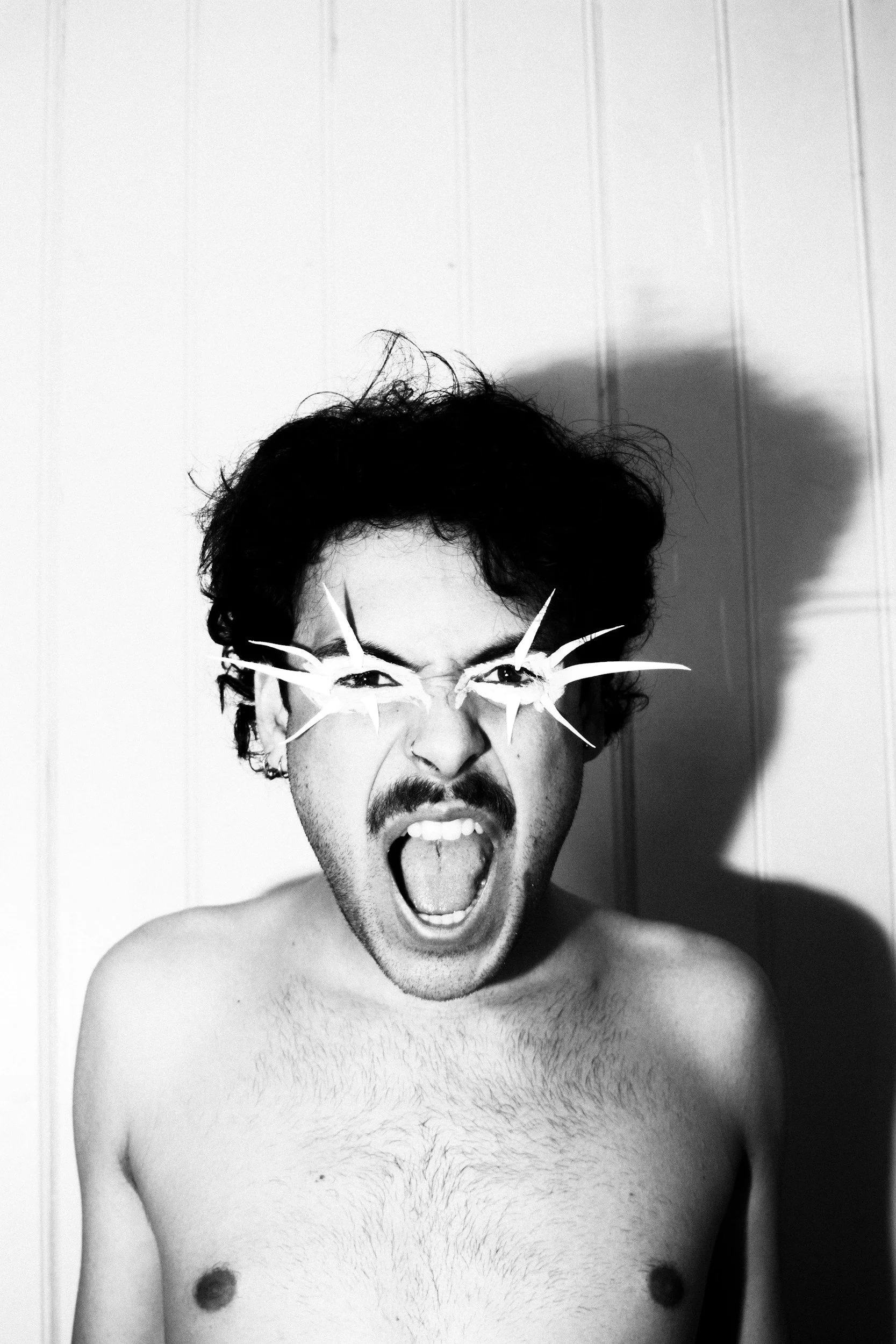 A shirtless man with messy hair and a mustache wearing glasses made of star-shaped paper cuts, shouting or yelling with an intense expression, in black and white.