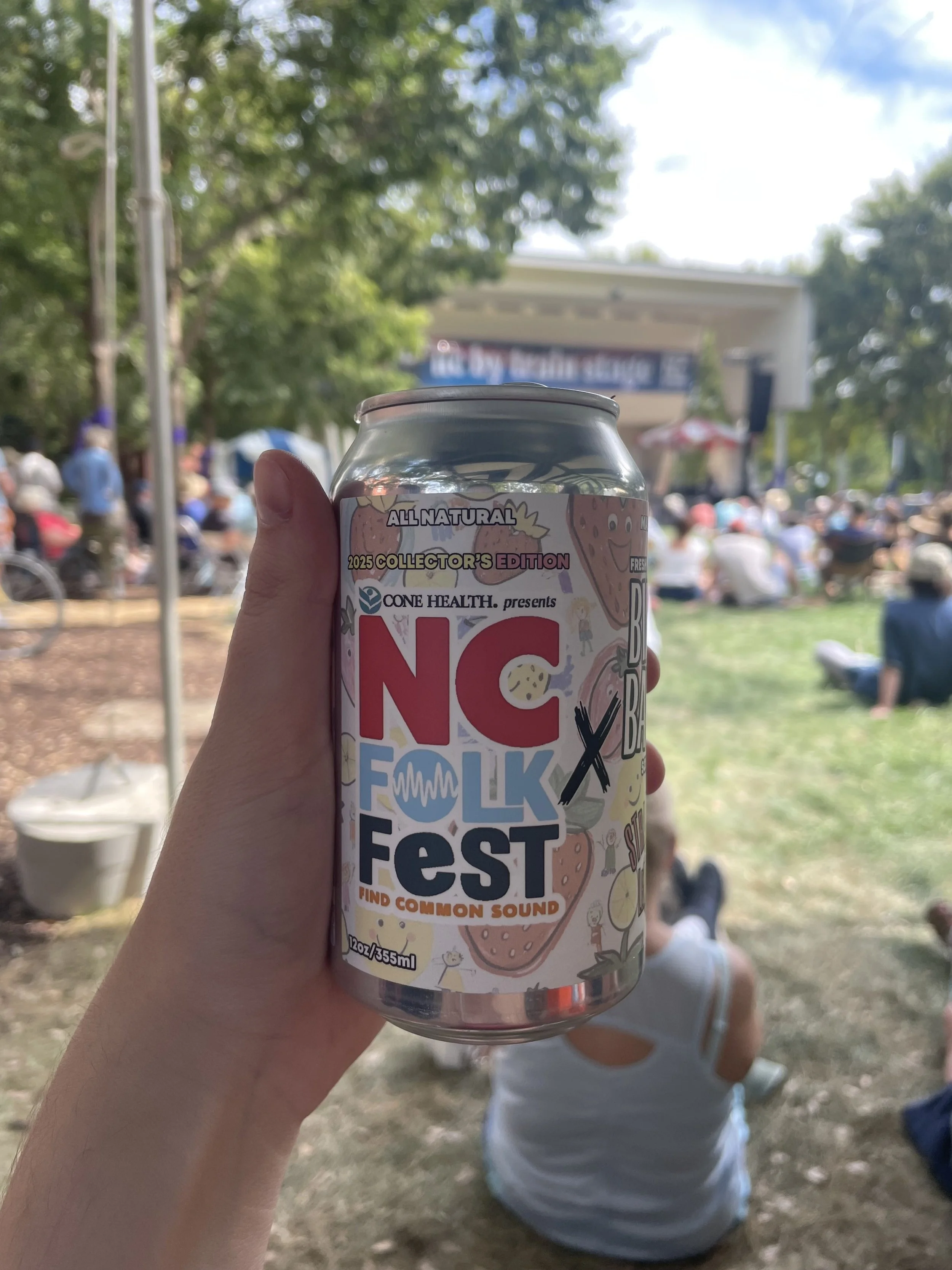 Hand holding a can of NC Folk Fest beverage with a blurred outdoor festival scene in the background