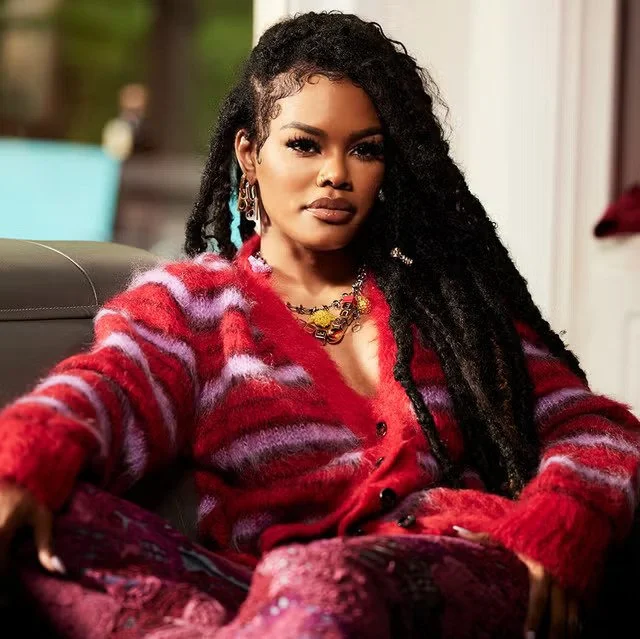 Teyana Taylor sitting indoors, wearing a colorful red and purple fuzzy sweater, multiple necklaces, and earrings, with a serious expression.