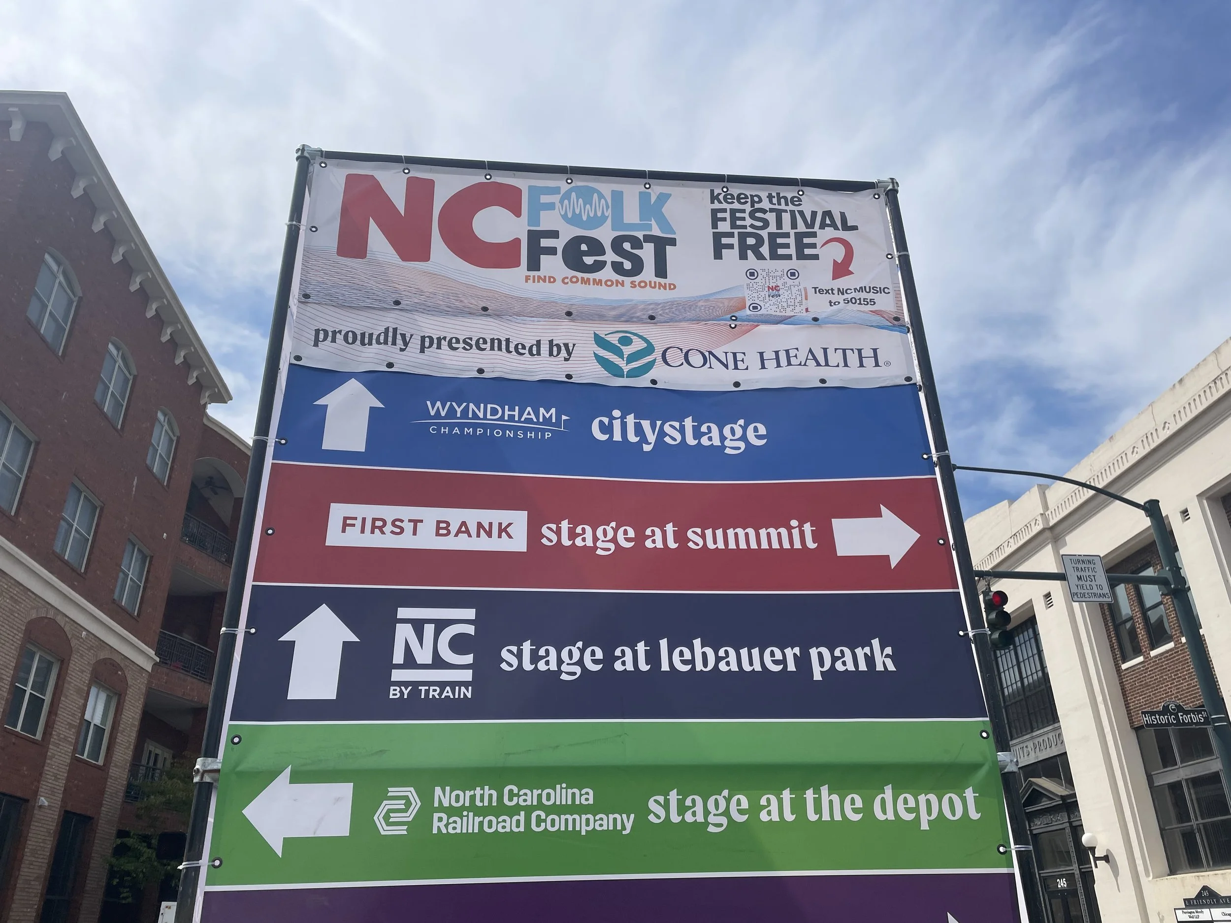 Directional signage for NC Folk Fest, listing stages at different locations, including at Summit, Lebauer Park, and Depot, with arrows pointing in various directions.