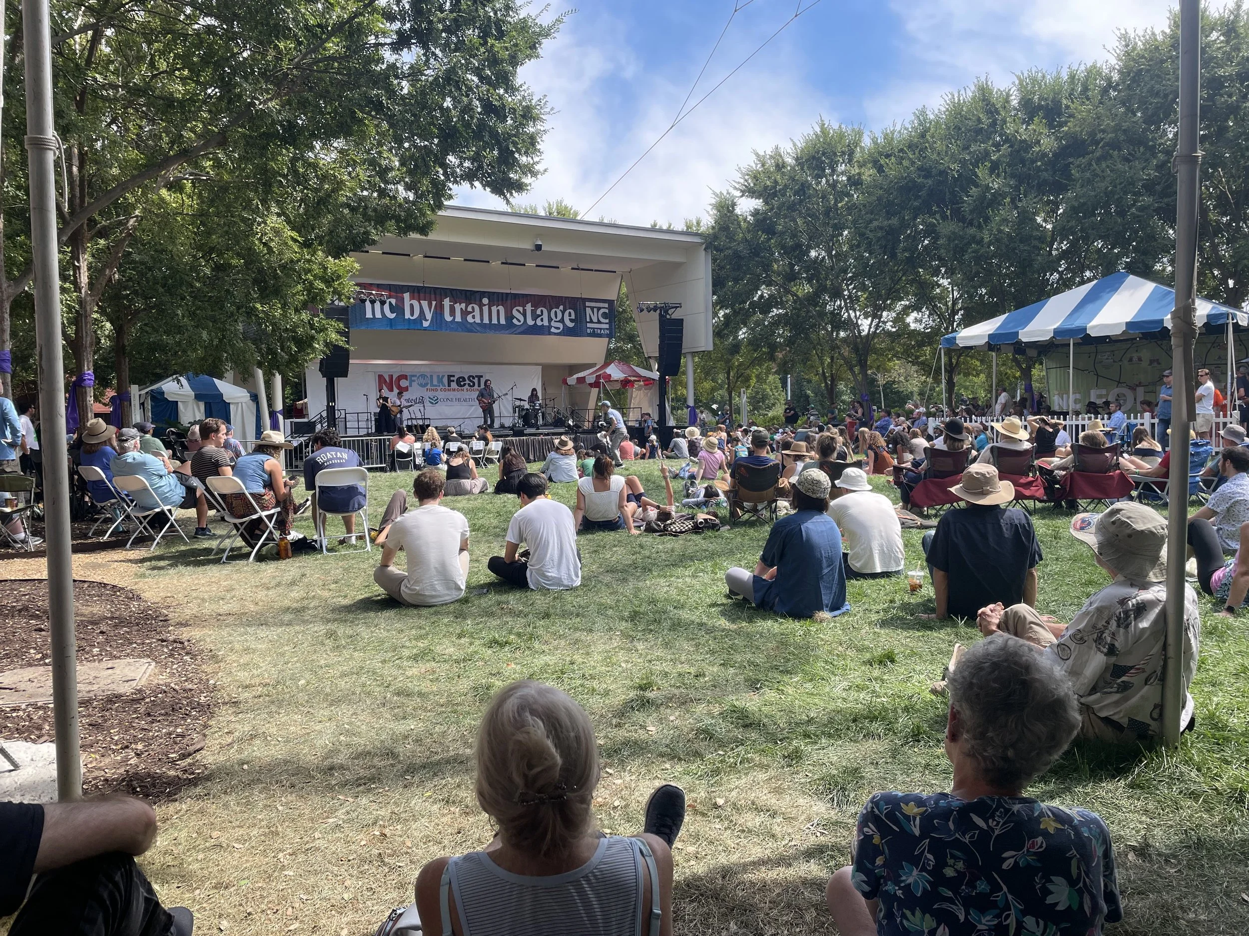 Outdoor concert festival with a stage, performers, and audience seated and standing on grass and chairs in a park, under trees on a sunny day.