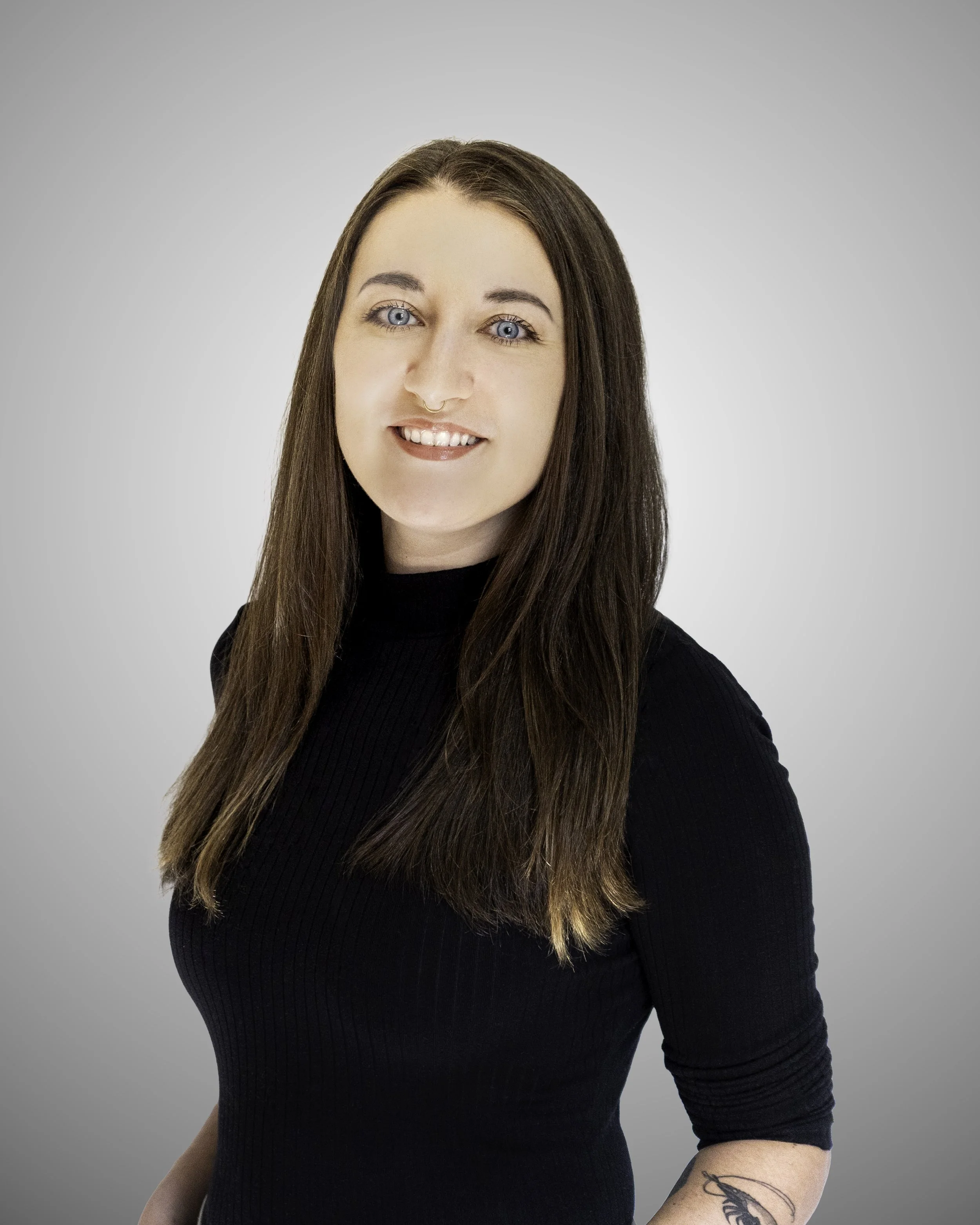 A young woman with long brown hair, blue eyes, wearing a black long-sleeve top, smiling, against a neutral gray background.