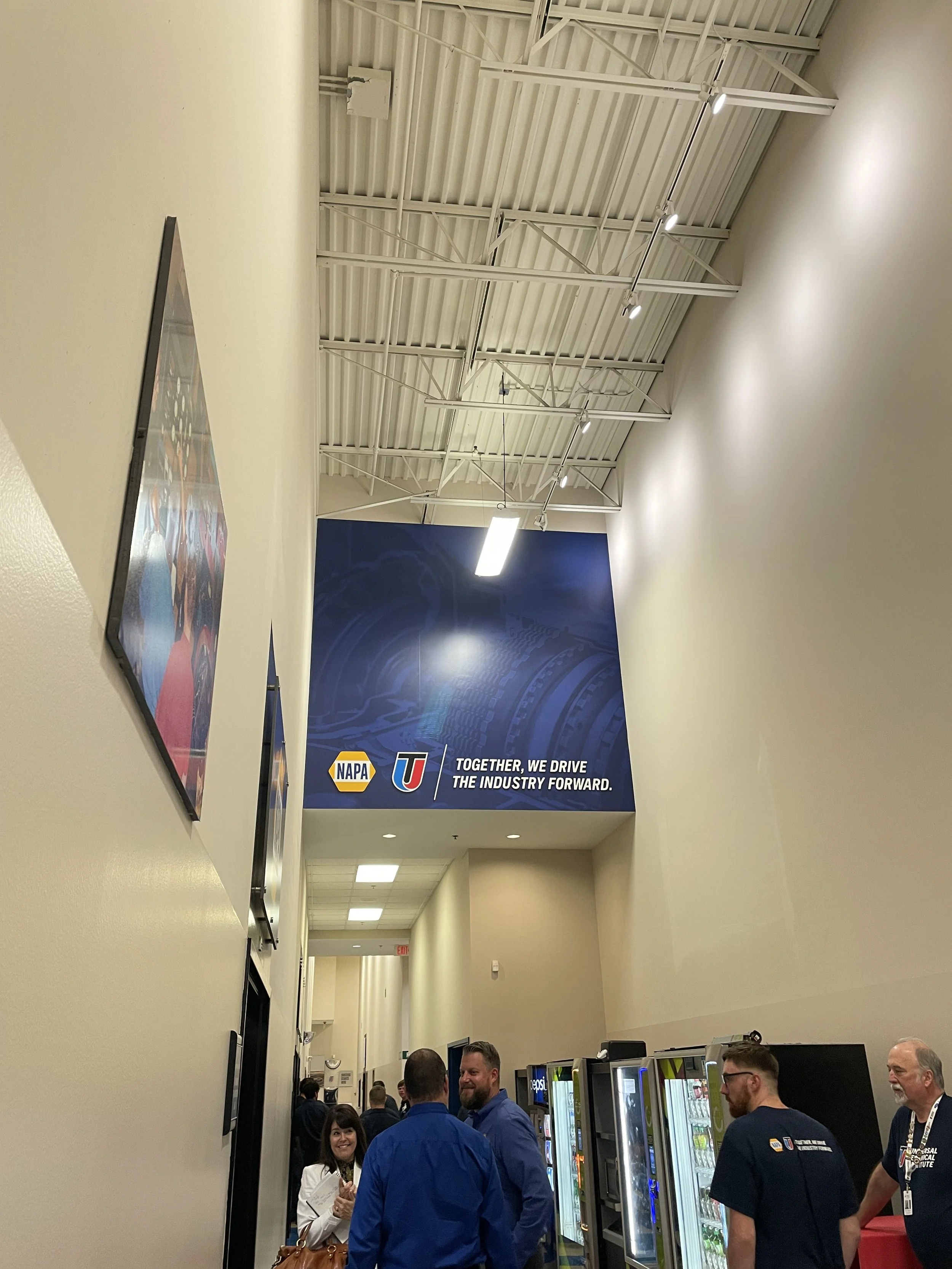 People standing in a hallway with vending machines and a large NAPA banner on the wall that reads "Together, we drive the industry forward."
