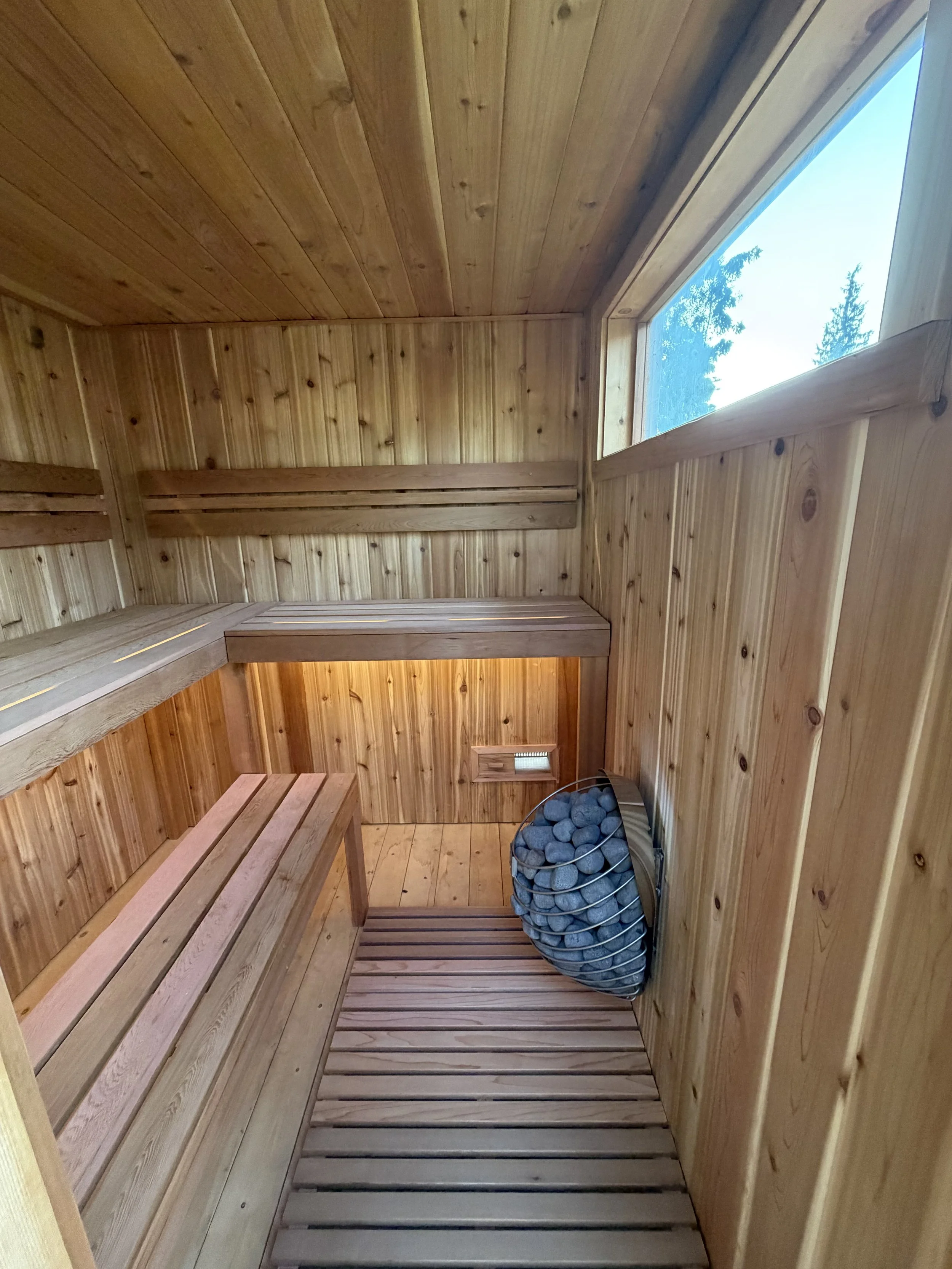 Wood-paneled indoor sauna with benches, a small window, and a heater filled with stones in a circular metal container.
